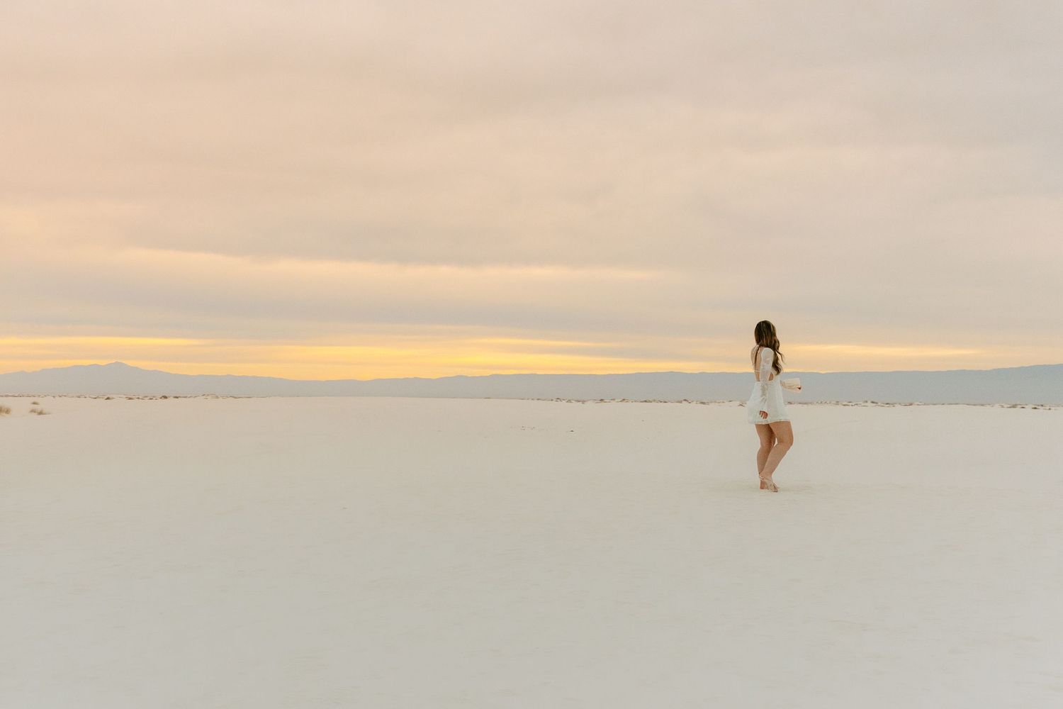 Woman in white mini dress stands alone on vast white sand dunes at colorful sunset.