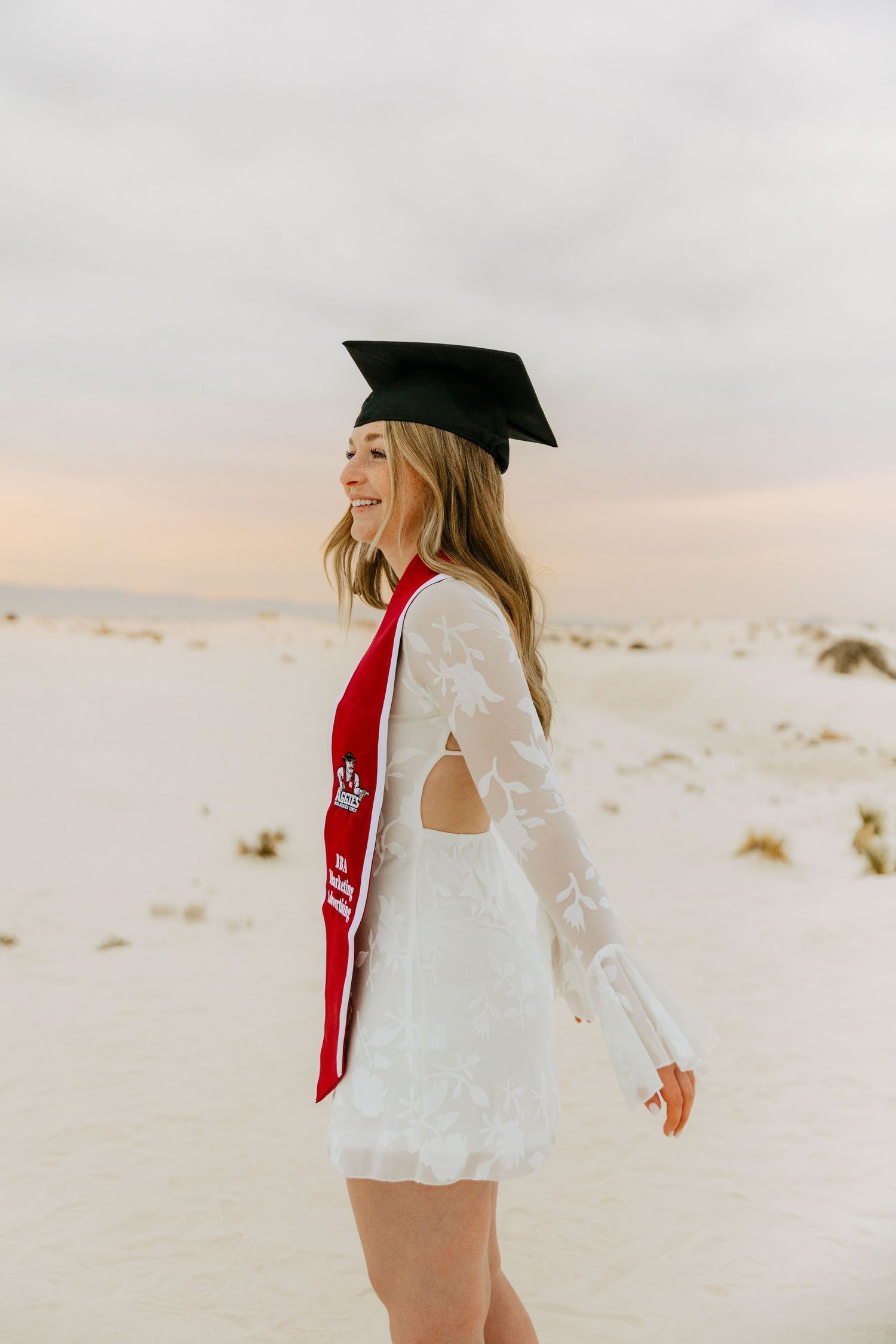 Smiling graduate in white lace mini dress with cap and red sash on white sand dunes.