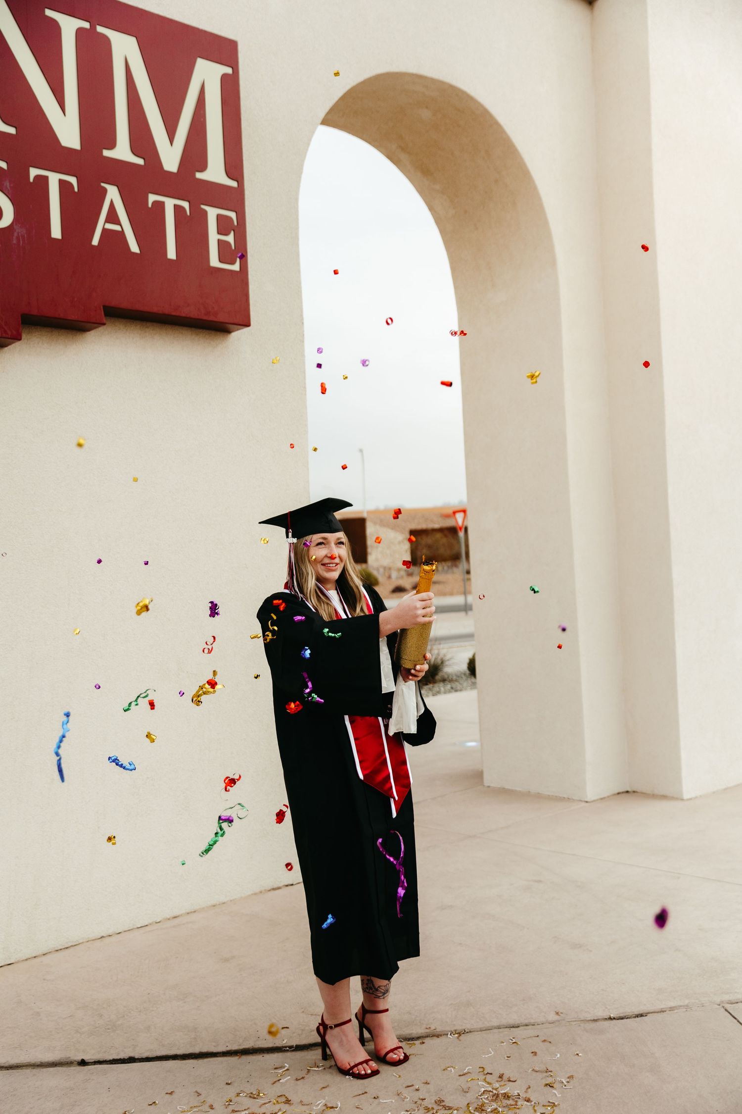 Smiling graduate in black cap and gown holding diploma with colorful confetti at NM State University entrance.