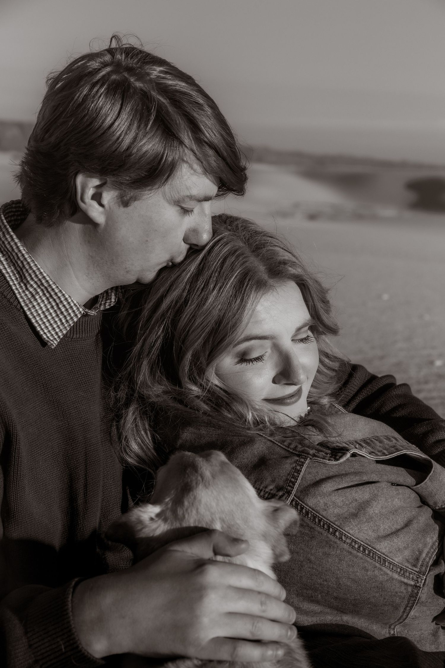 Black and white photo of a romantic couple cuddling with a small dog on sandy dunes.