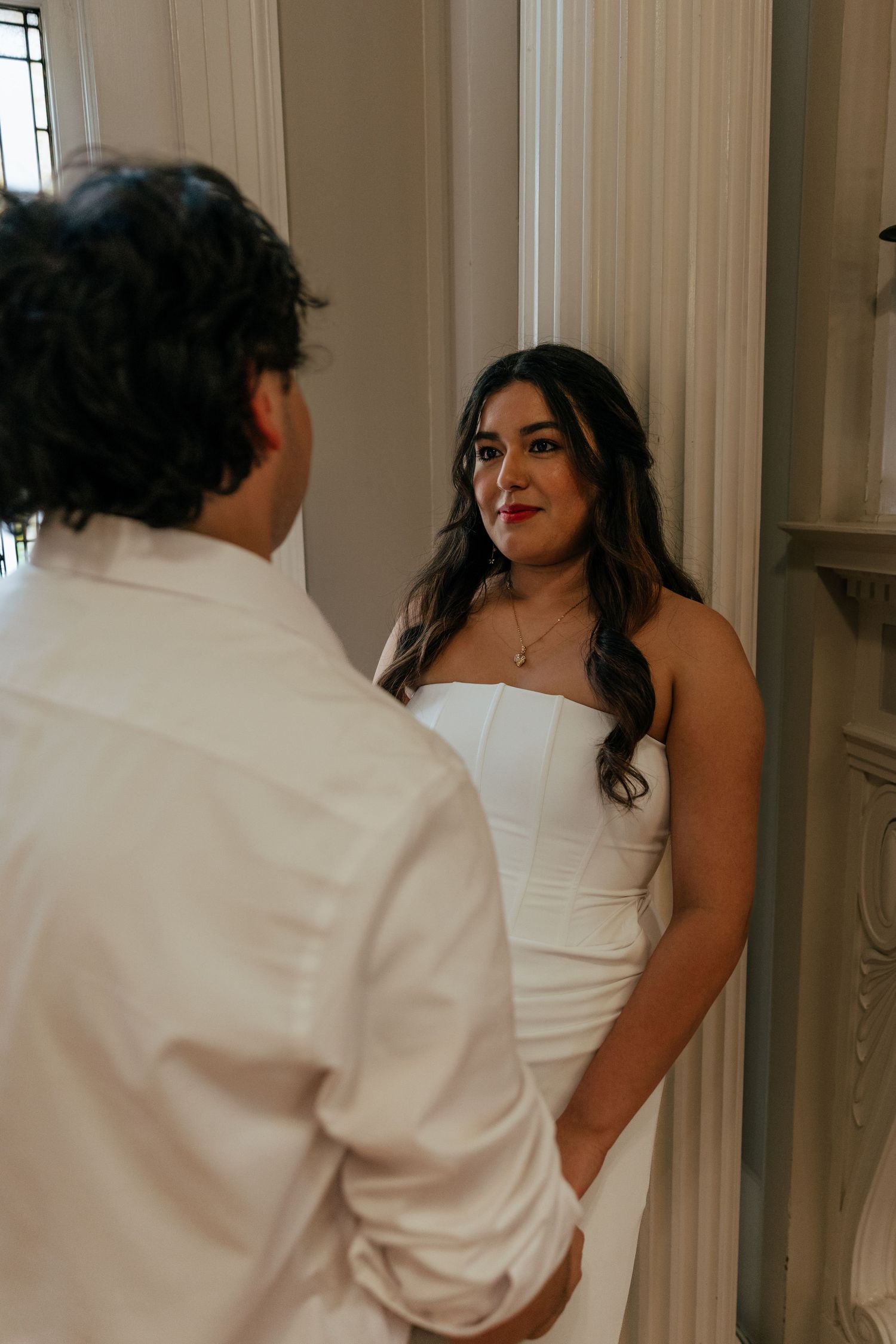 Bride in strapless white gown shares an intimate first look moment with groom in white shirt indoors.