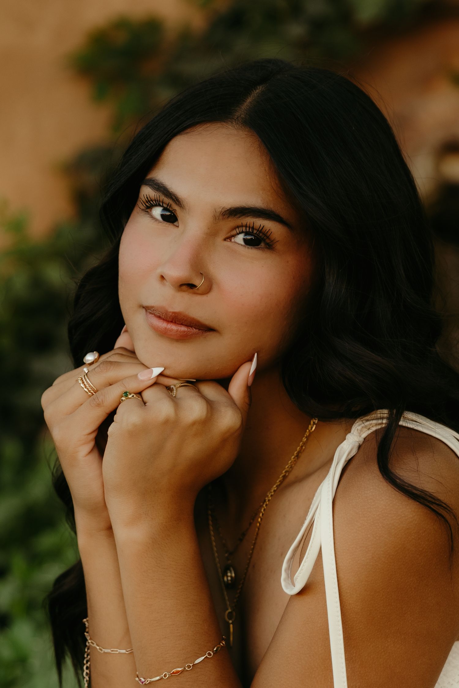 Close-up portrait of young Latina woman with gold jewelry and rings posing outdoors in white dress.
