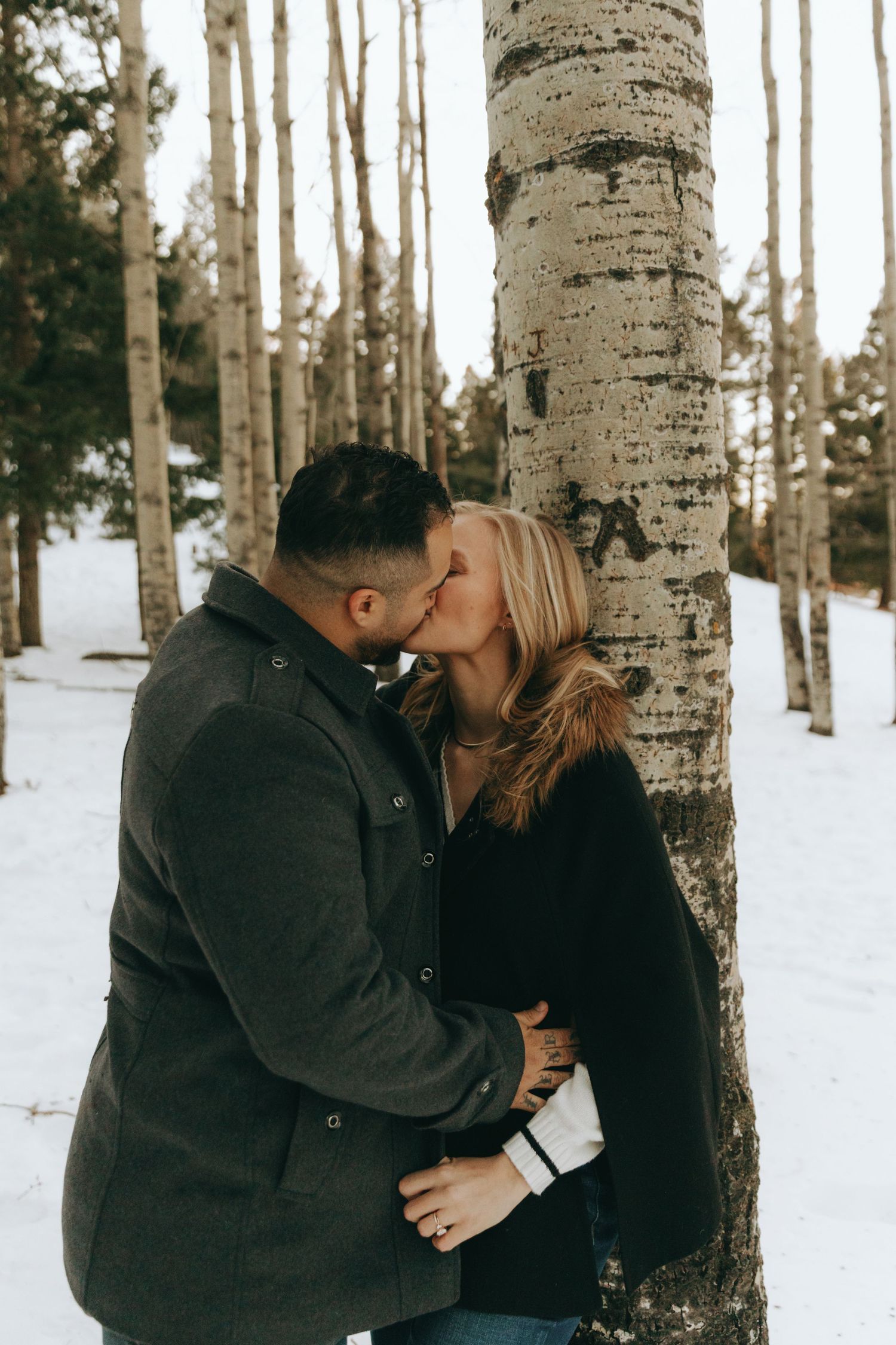 Couple kissing beside a birch tree in a snowy winter forest, wearing dark coats in a romantic outdoor engagement photo.