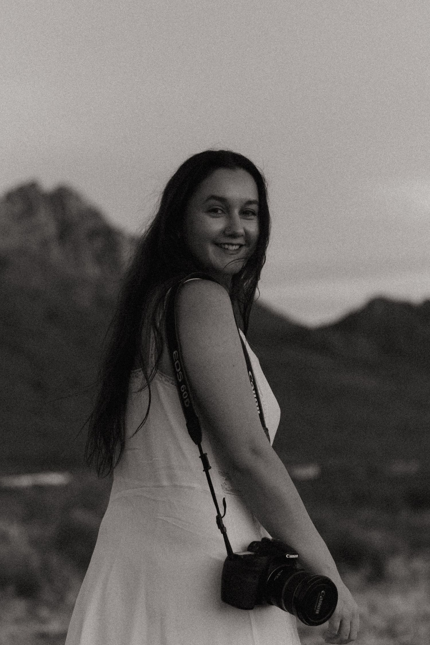 Black and white portrait of smiling woman with Canon camera and mountain backdrop.