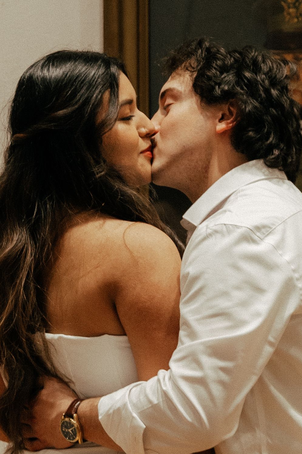 A couple sharing a romantic kiss, woman in white strapless dress with long dark hair, man in white shirt.