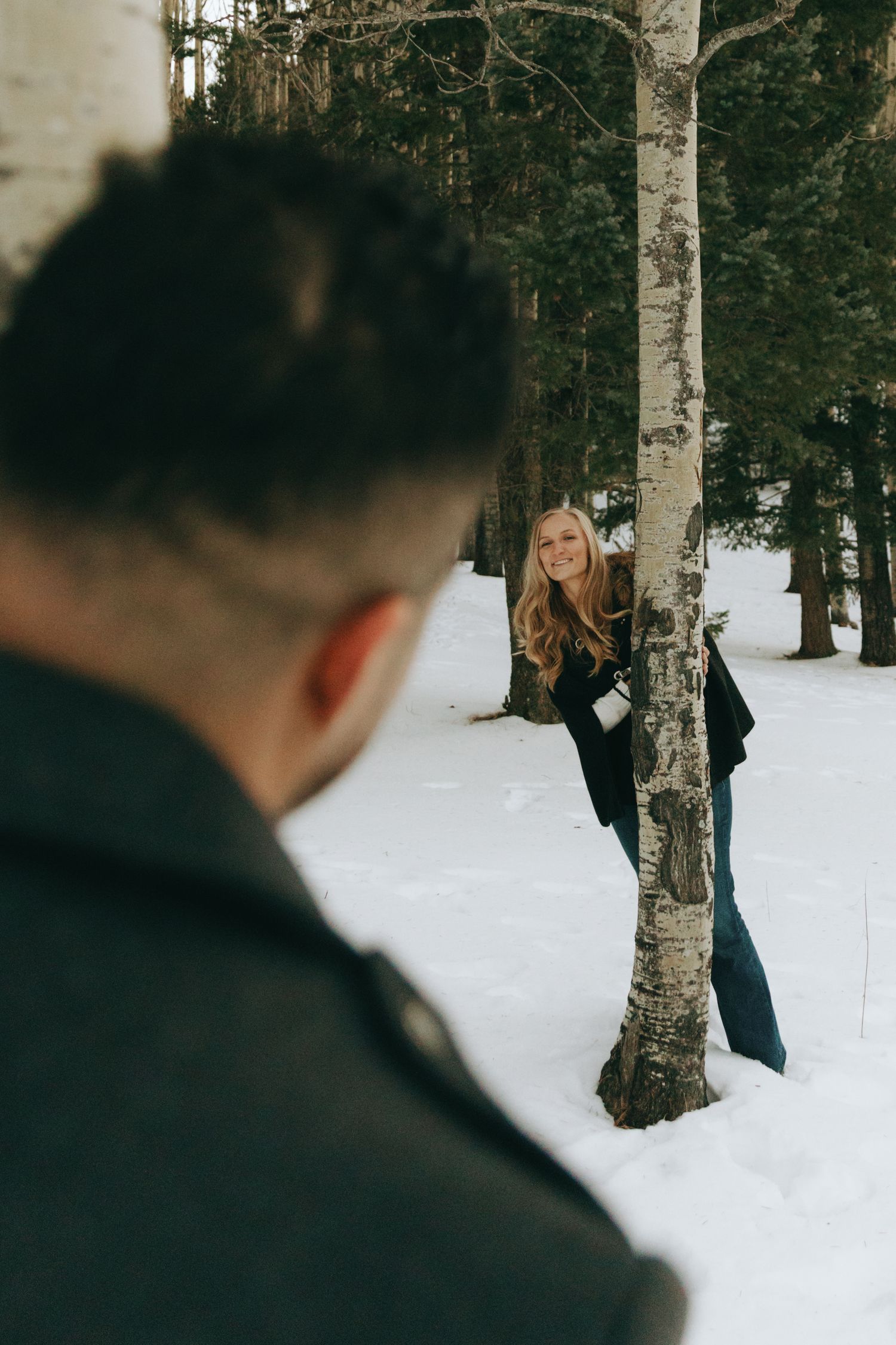 A smiling woman in jeans leans against a birch tree in a snowy forest while a man watches her from the foreground.