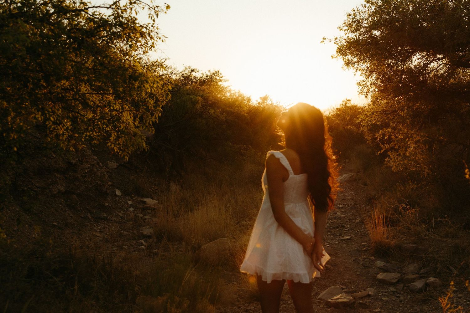 Young woman in white dress standing on a sunlit forest path during golden hour sunset.