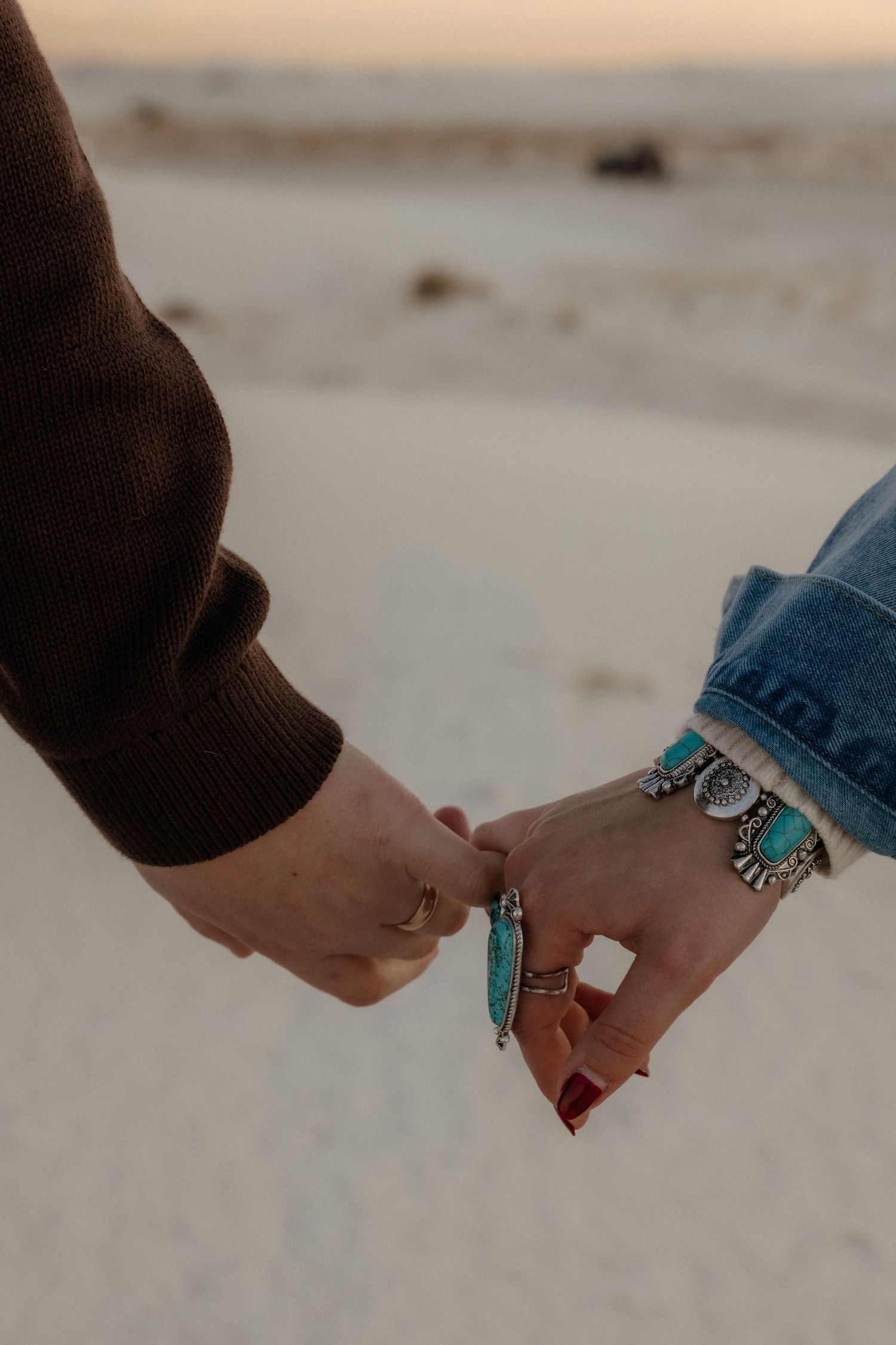 Close-up of couple holding hands on sandy beach, woman wearing turquoise silver jewelry and rings at sunset.