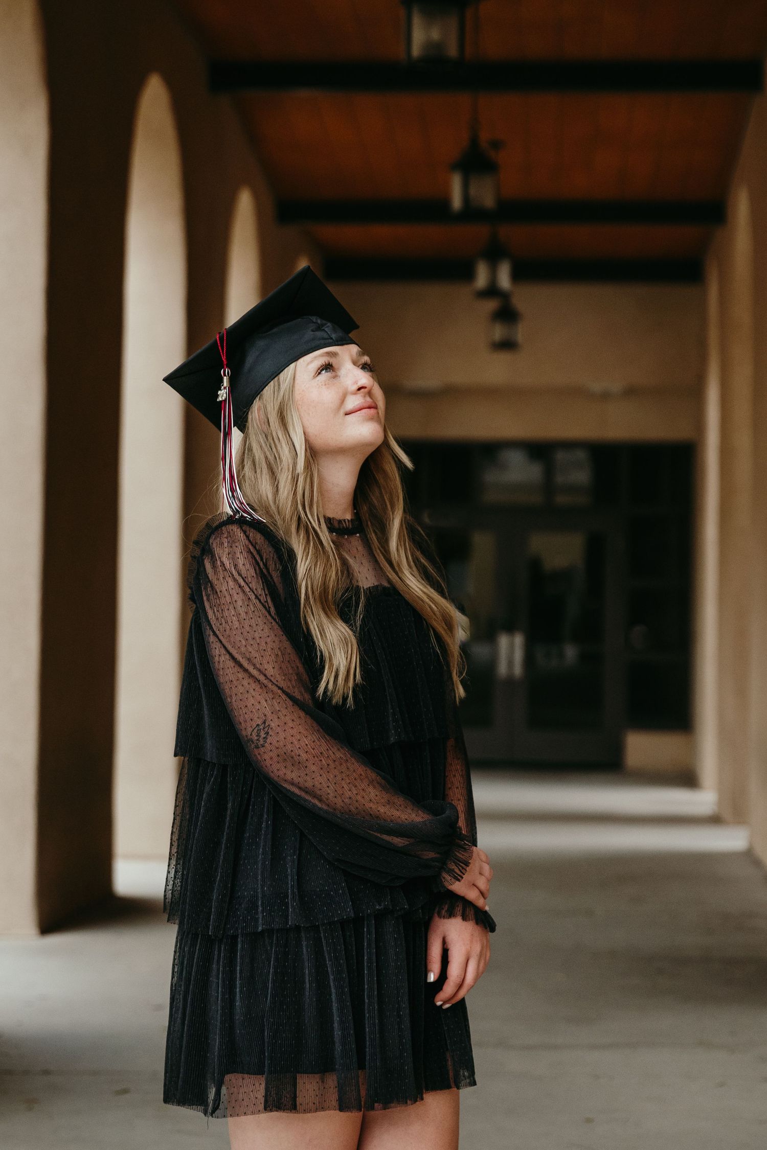 Graduate in black cap and sheer dress gazing upward in arched campus corridor.