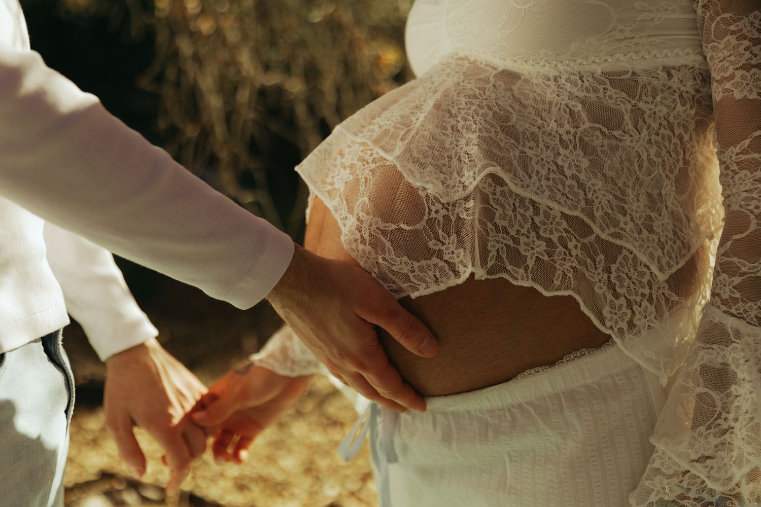 Pregnant bride in lace wedding dress with partner touching baby bump outdoors.