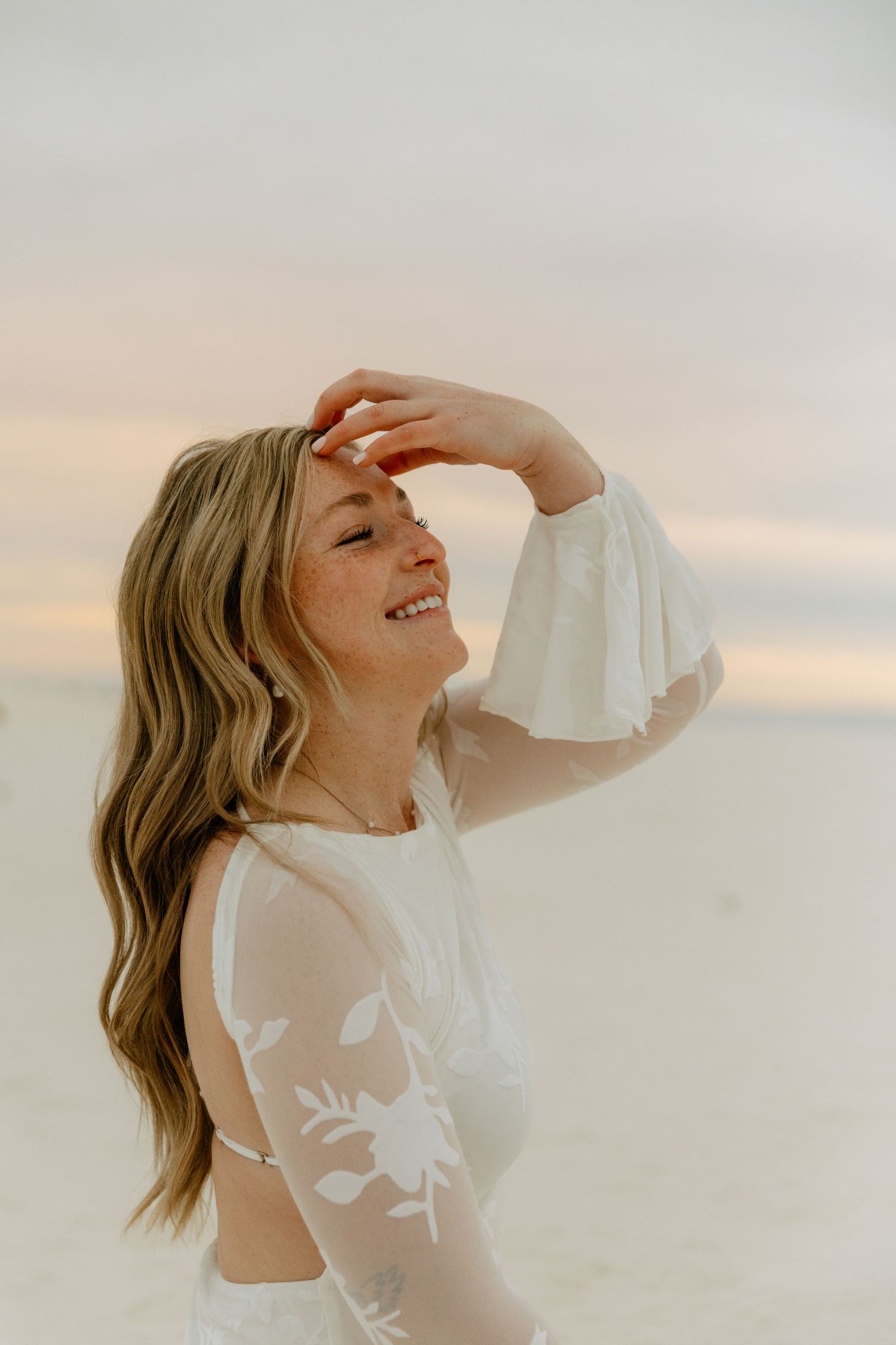 Joyful blonde woman in white lace dress laughs with hand raised to forehead on sunny beach.