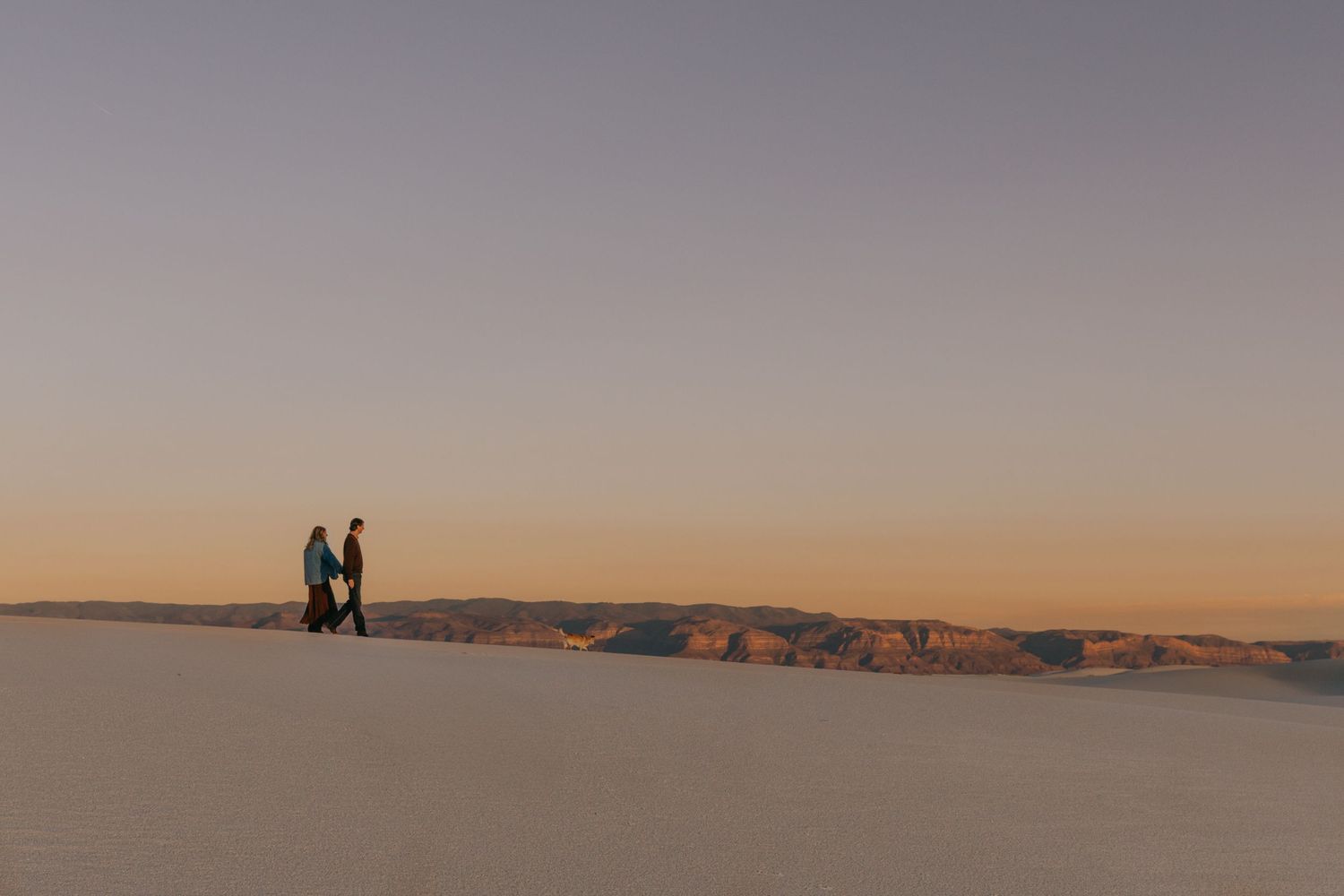 Couple holding hands while walking along white sand dunes at sunset with mountains.