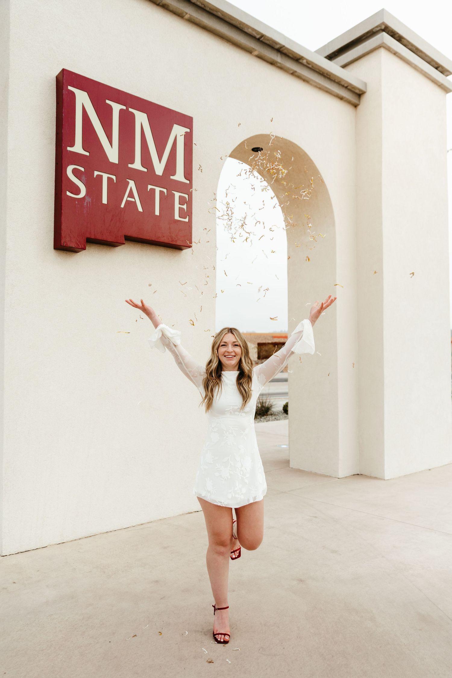 Woman in white dress celebrating graduation at NM State University sign while tossing confetti.