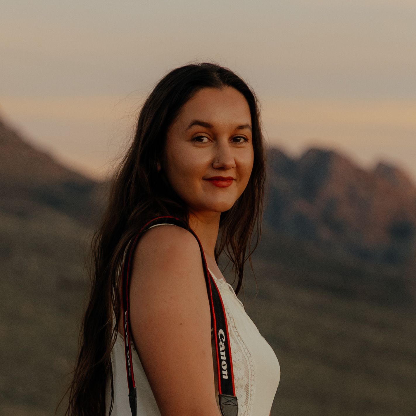 Young woman with long dark hair and Canon camera smiles at golden hour in mountains.