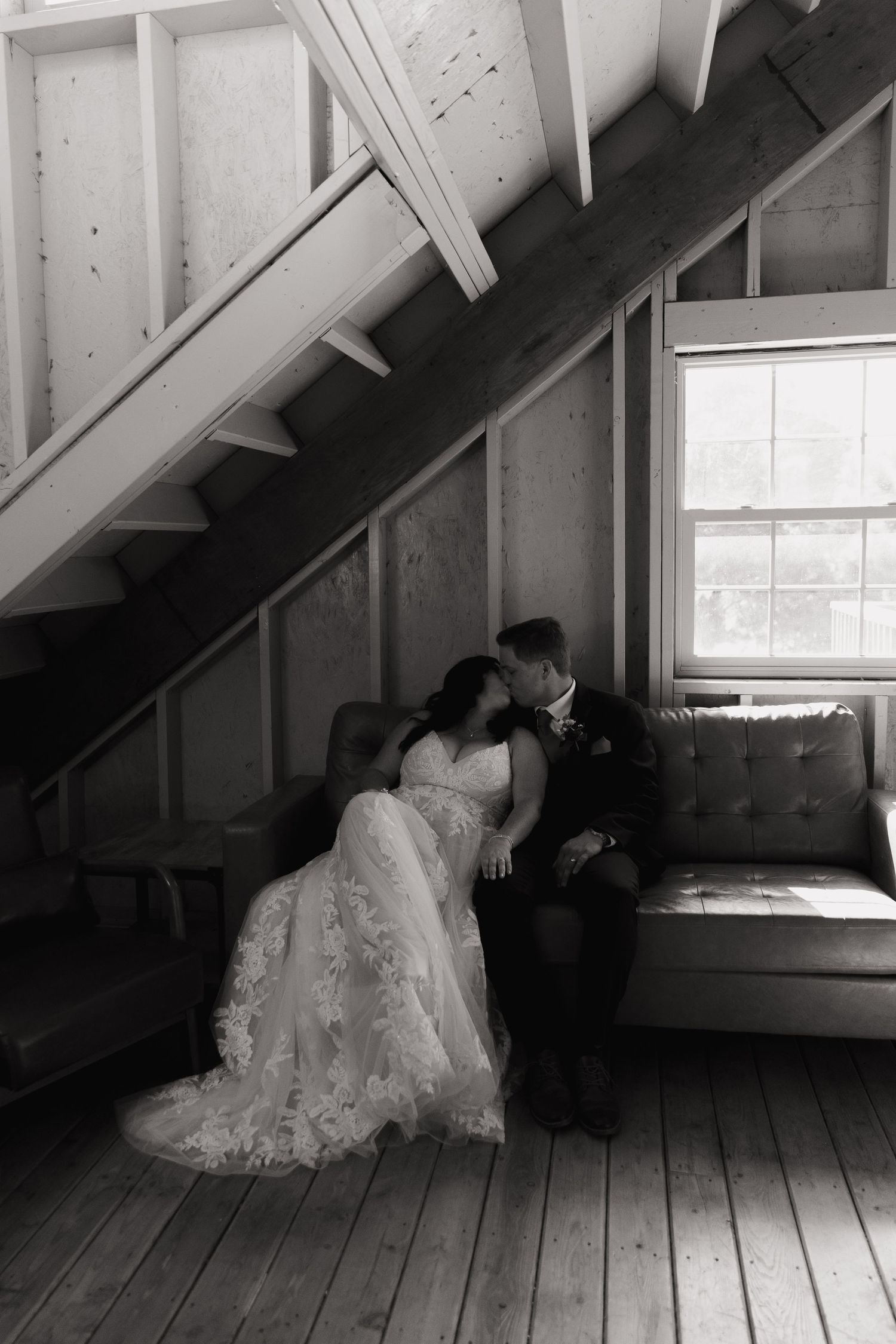 Black and white photo of a bride and groom sharing a kiss on a couch beneath a rustic staircase near a window.