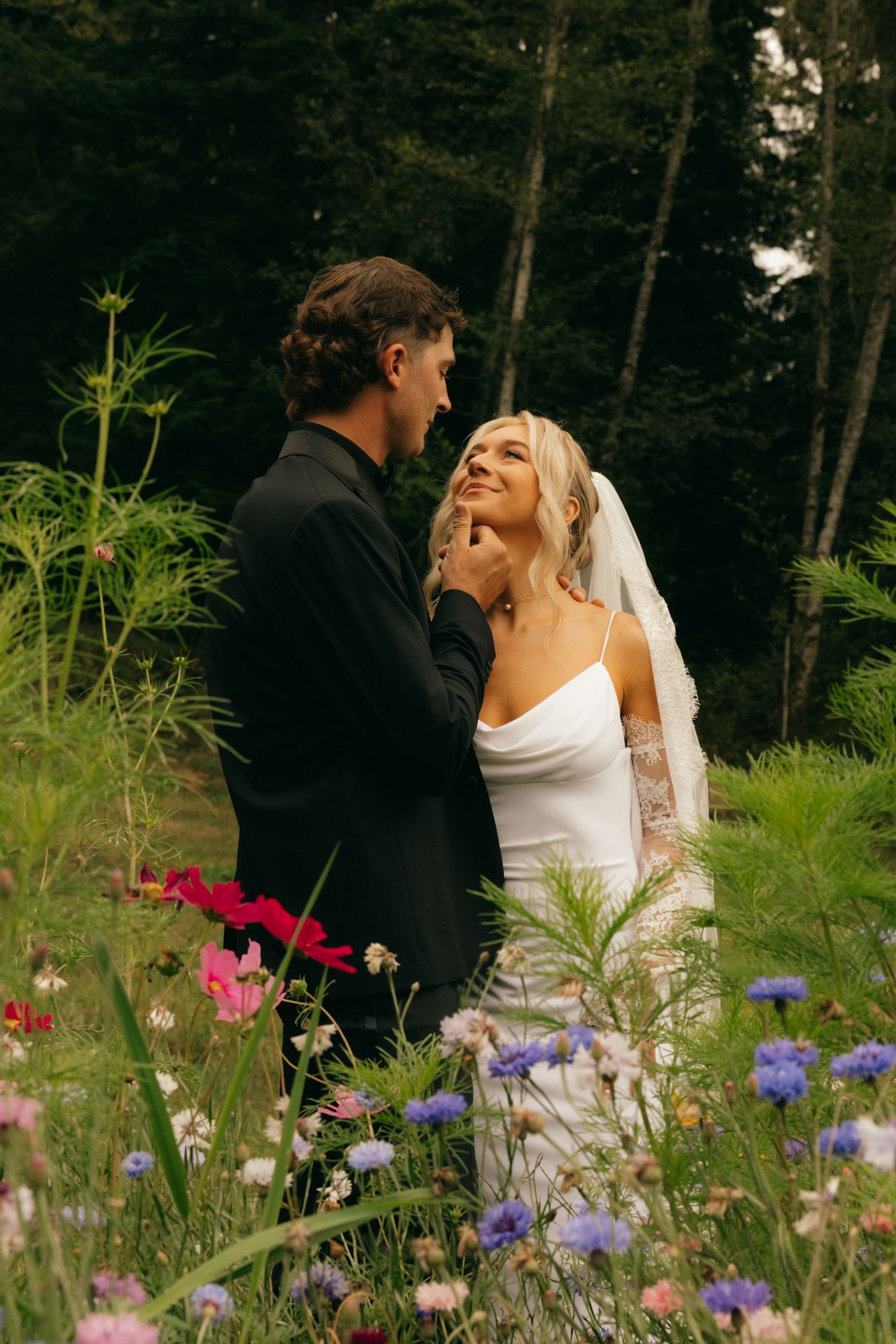 Bride and groom embrace among wildflowers in a forest setting, bride wearing white gown and veil.