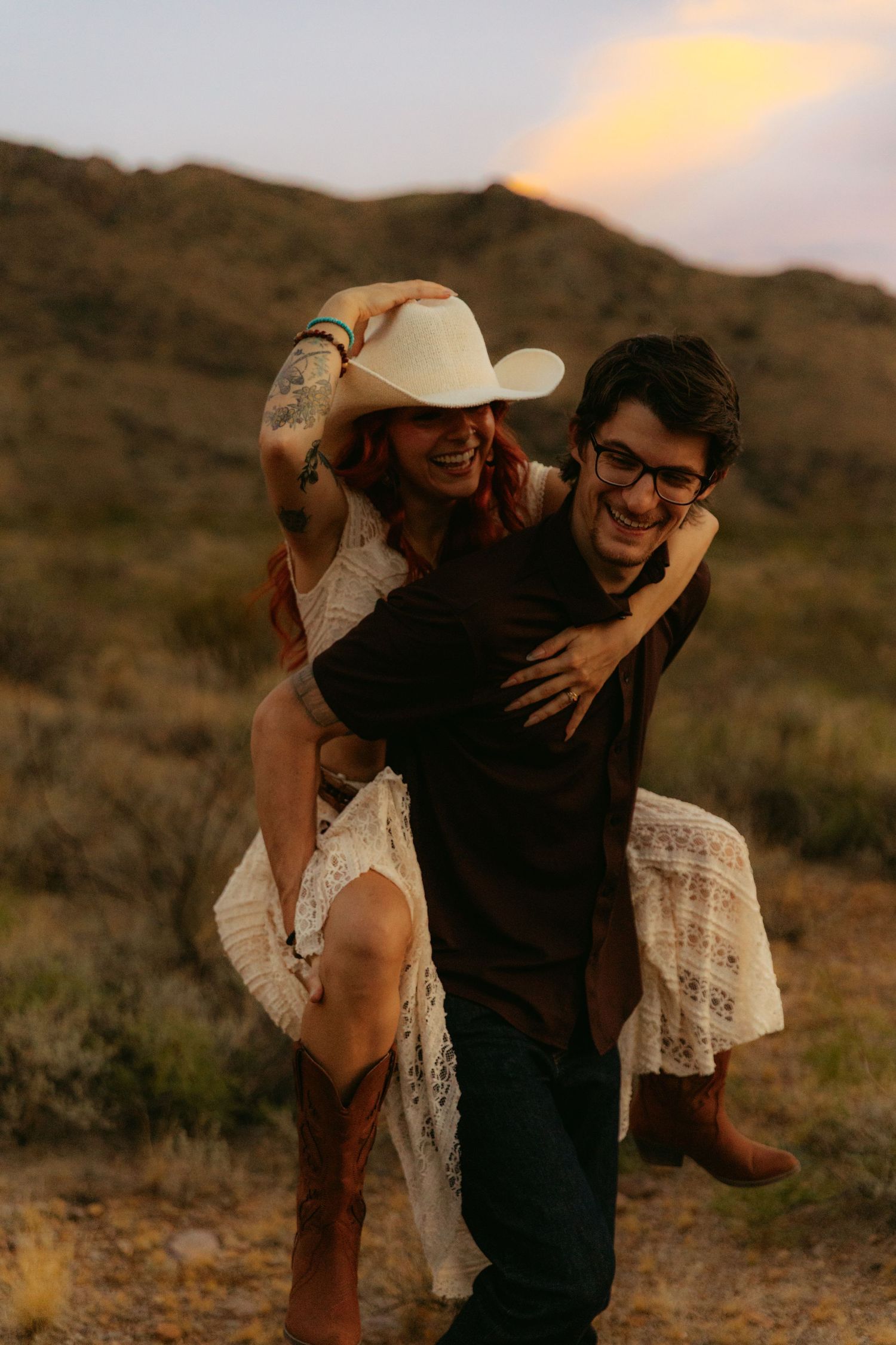 A joyful couple in desert landscape at sunset, man giving piggyback ride to woman in cowboy hat and lace dress.