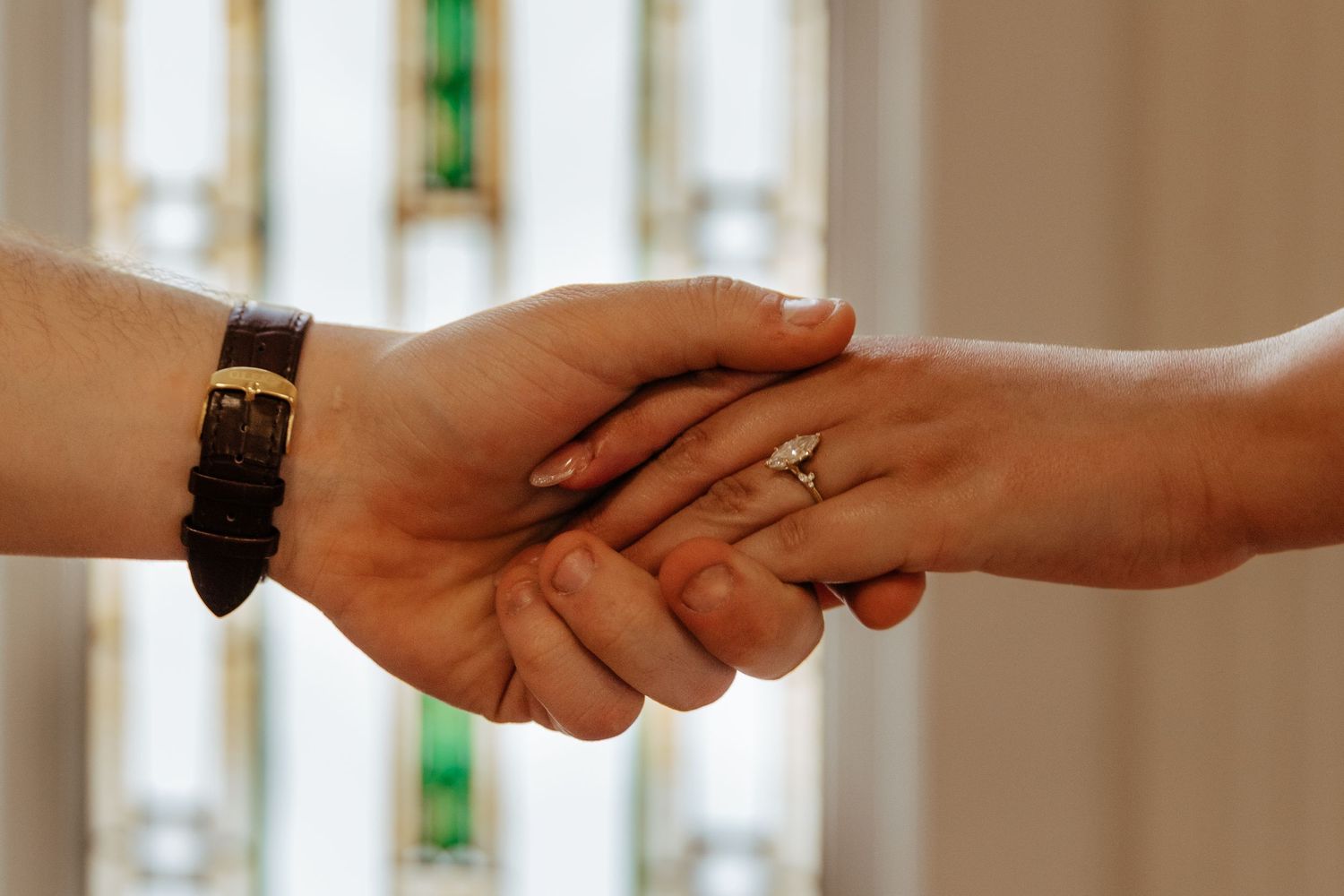 Two people shaking hands in a church, one wearing a watch and the other a diamond ring, with stained glass windows behind.