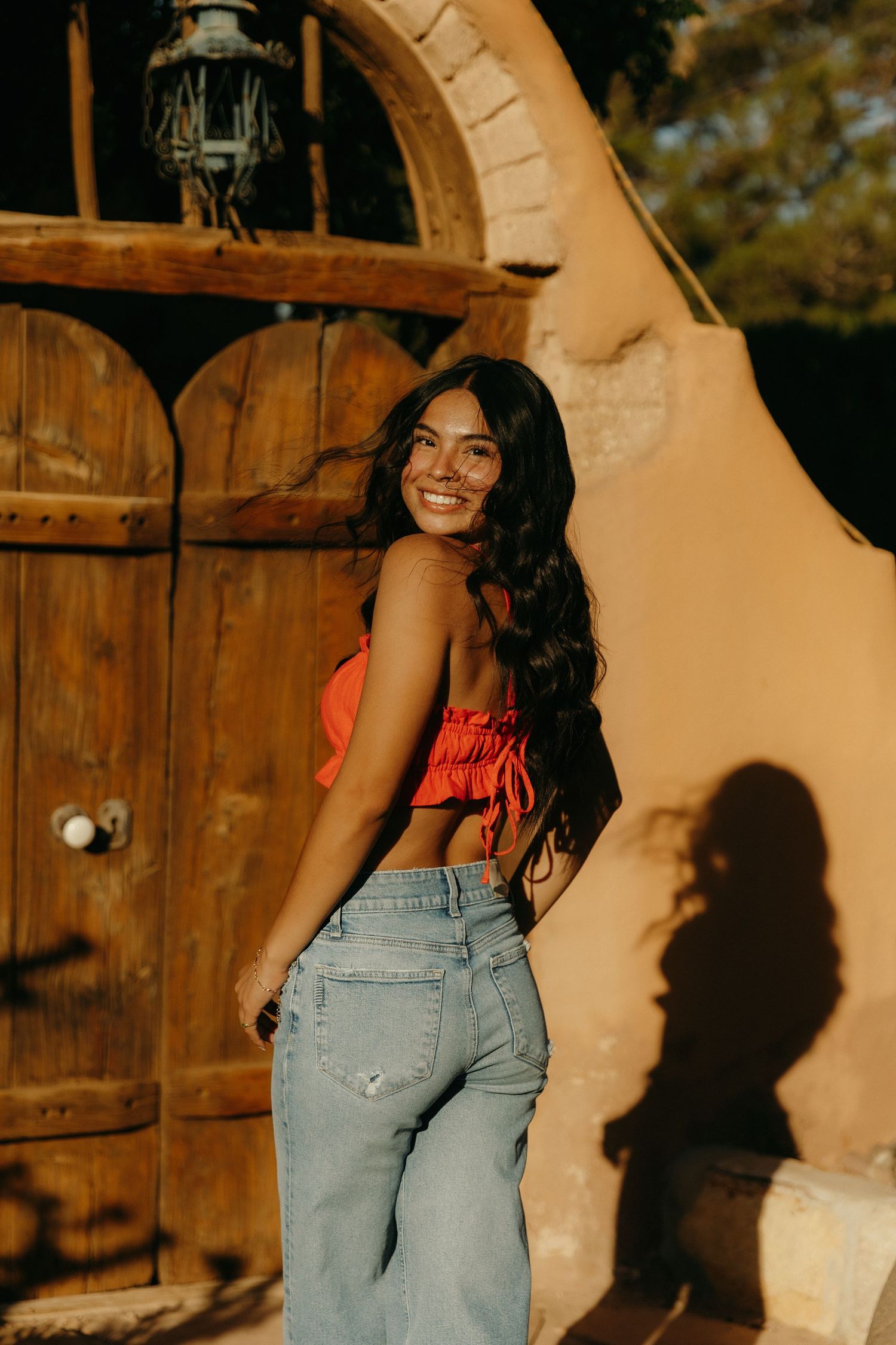 Young woman in red crop top and jeans smiling over shoulder near rustic wooden arched door in golden sunlight.