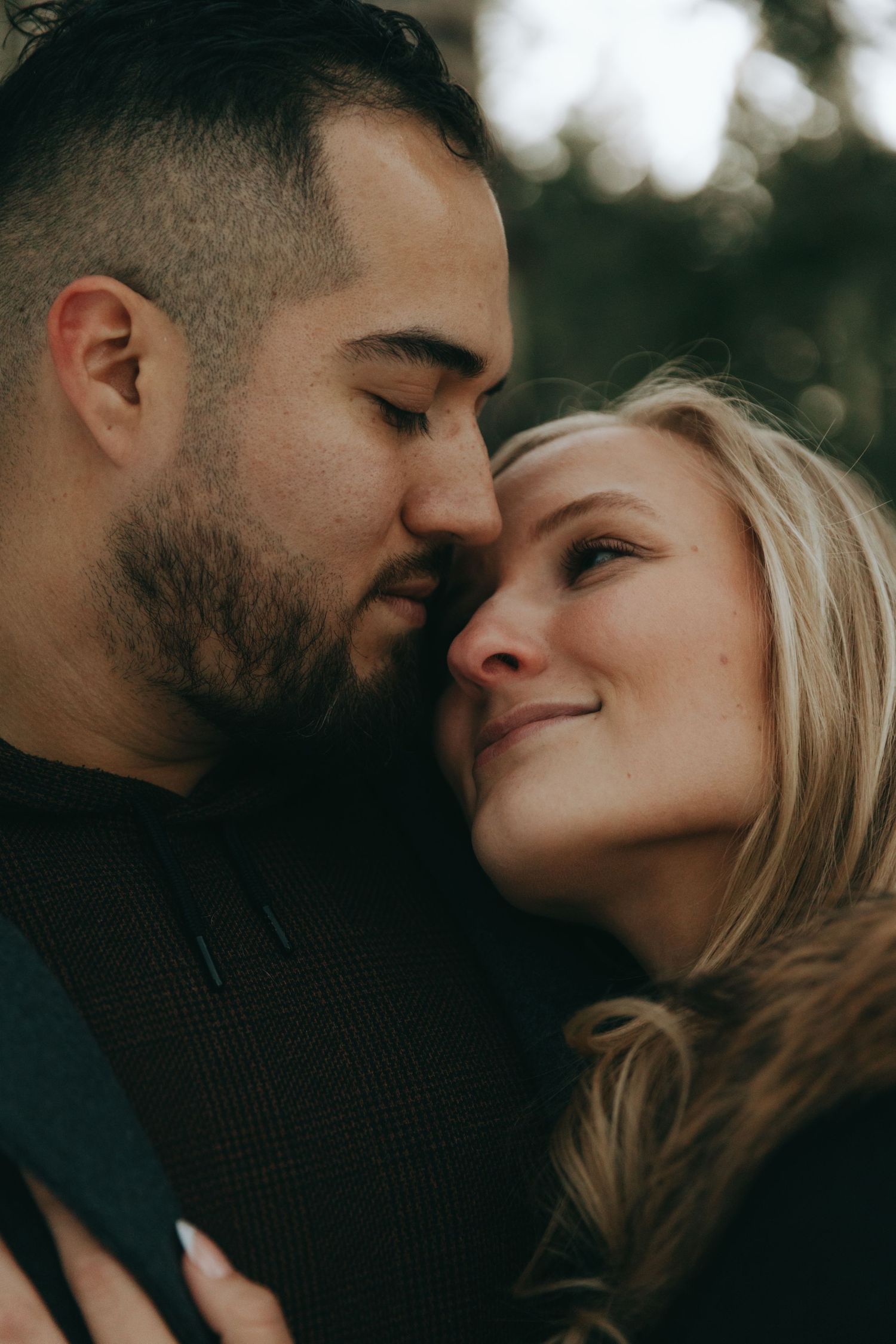 A couple shares an intimate moment, man with beard gently nuzzling smiling blonde woman outdoors.