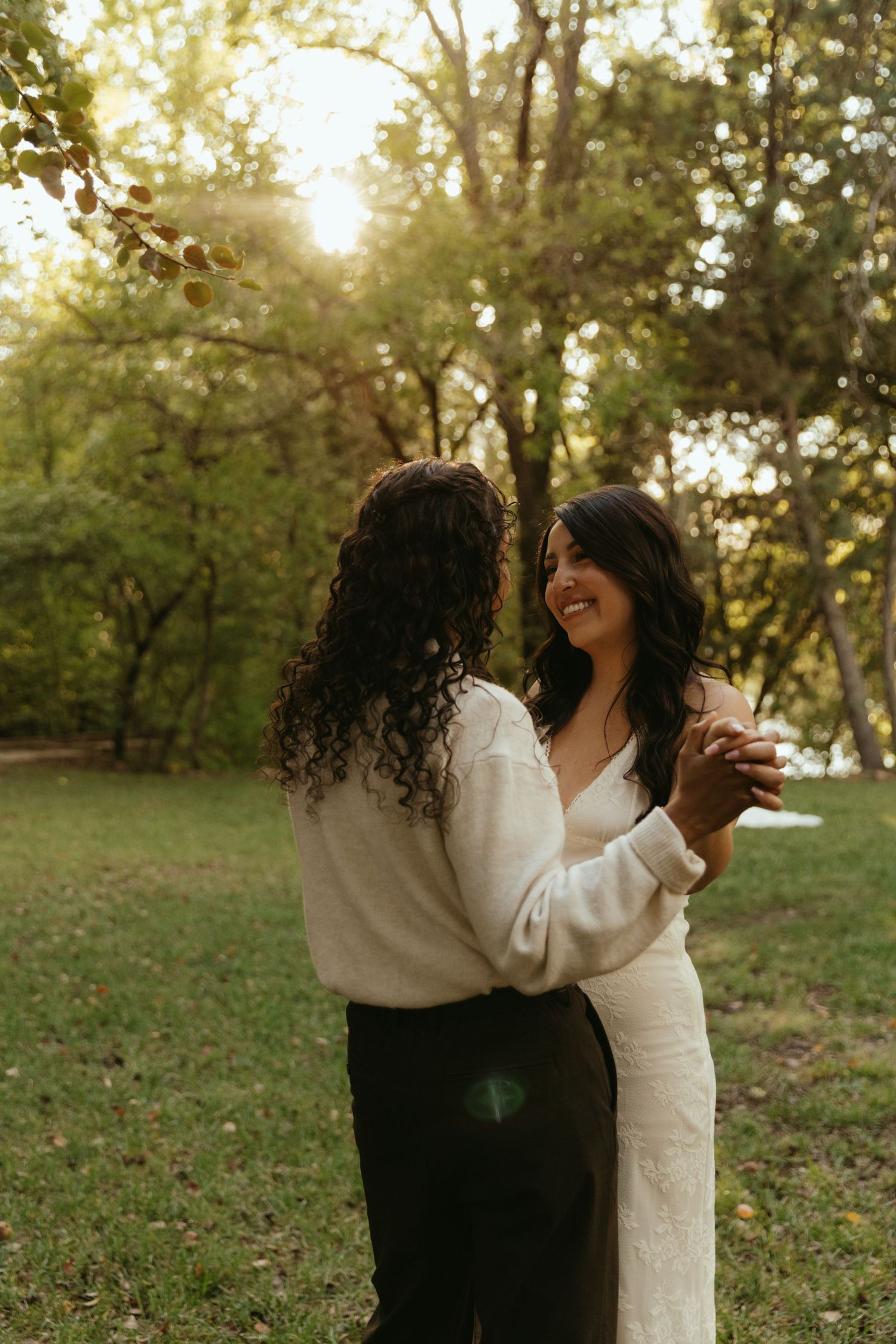 Two women with long dark curly hair hold hands and smile while dancing outdoors in a sunlit park surrounded by green trees.