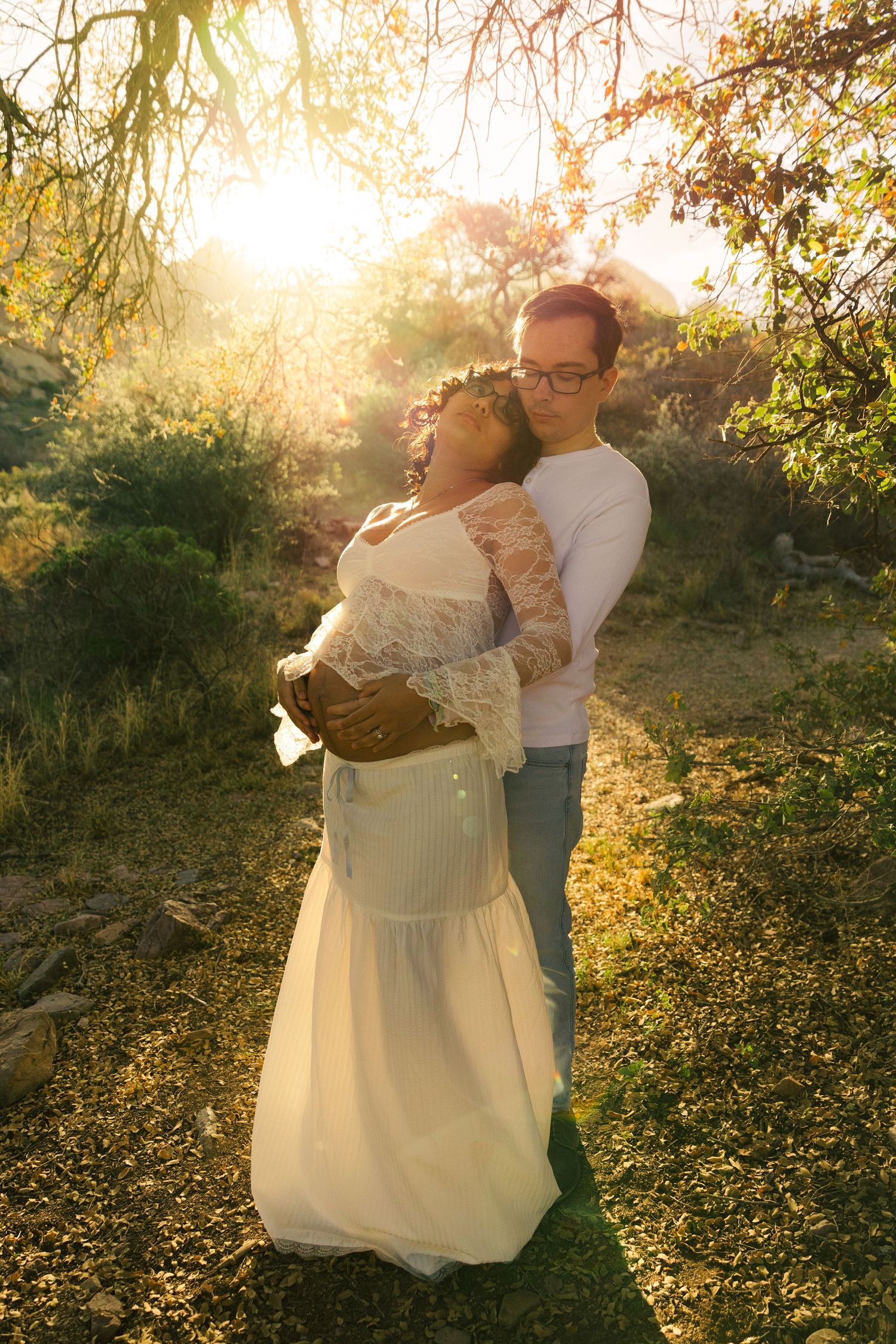 Expecting couple embracing in warm golden sunlight outdoors, woman in white lace maternity dress leaning back into partners arms.