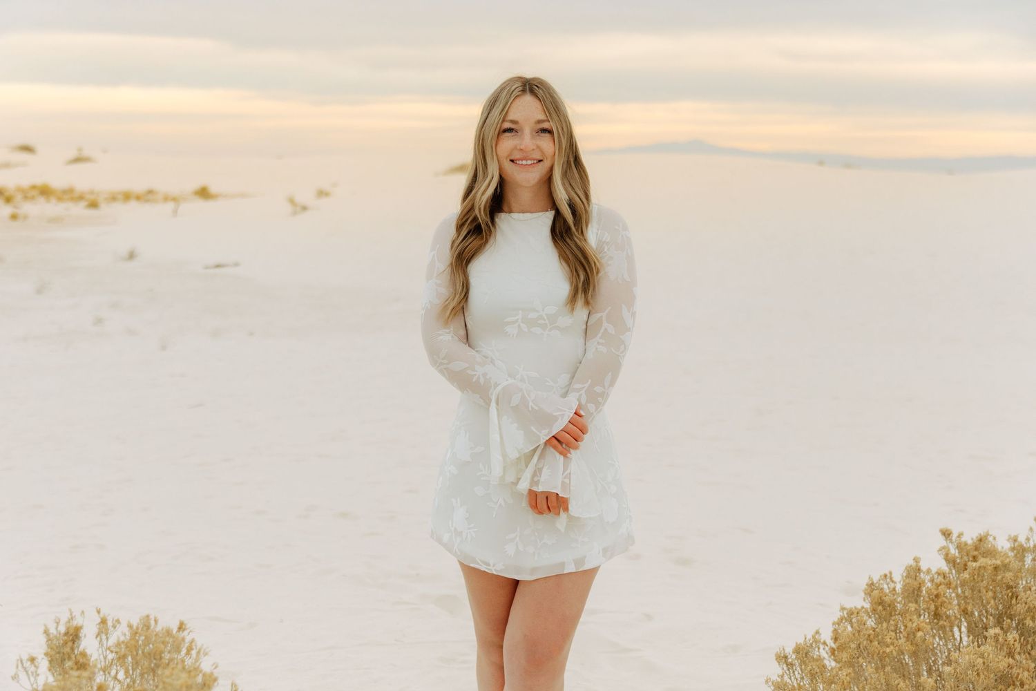 Smiling woman in white lace long-sleeve mini dress at White Sands dunes at sunset.