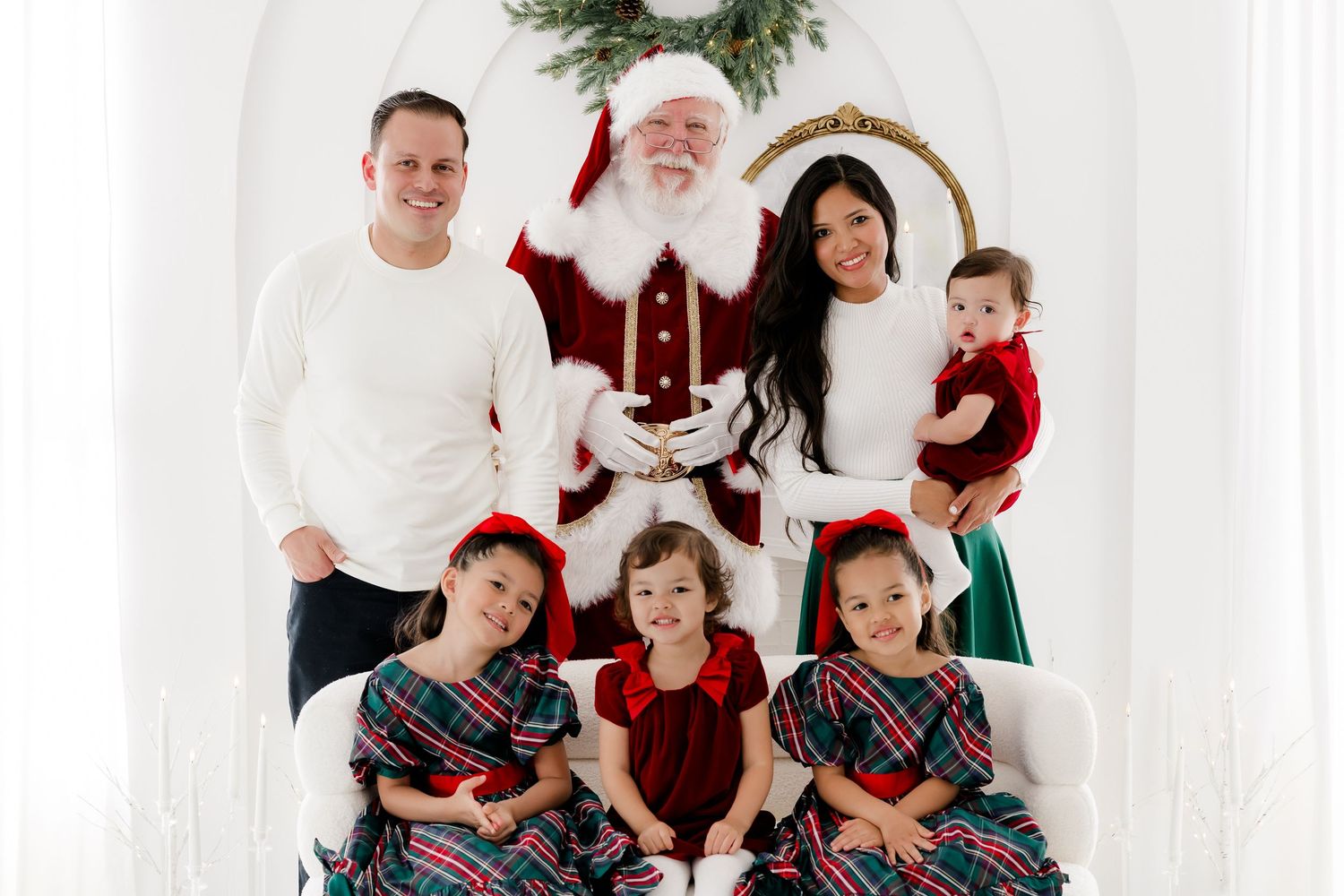 Family Christmas photo with Santa Claus, two parents, and four young children in festive holiday outfits in white studio.