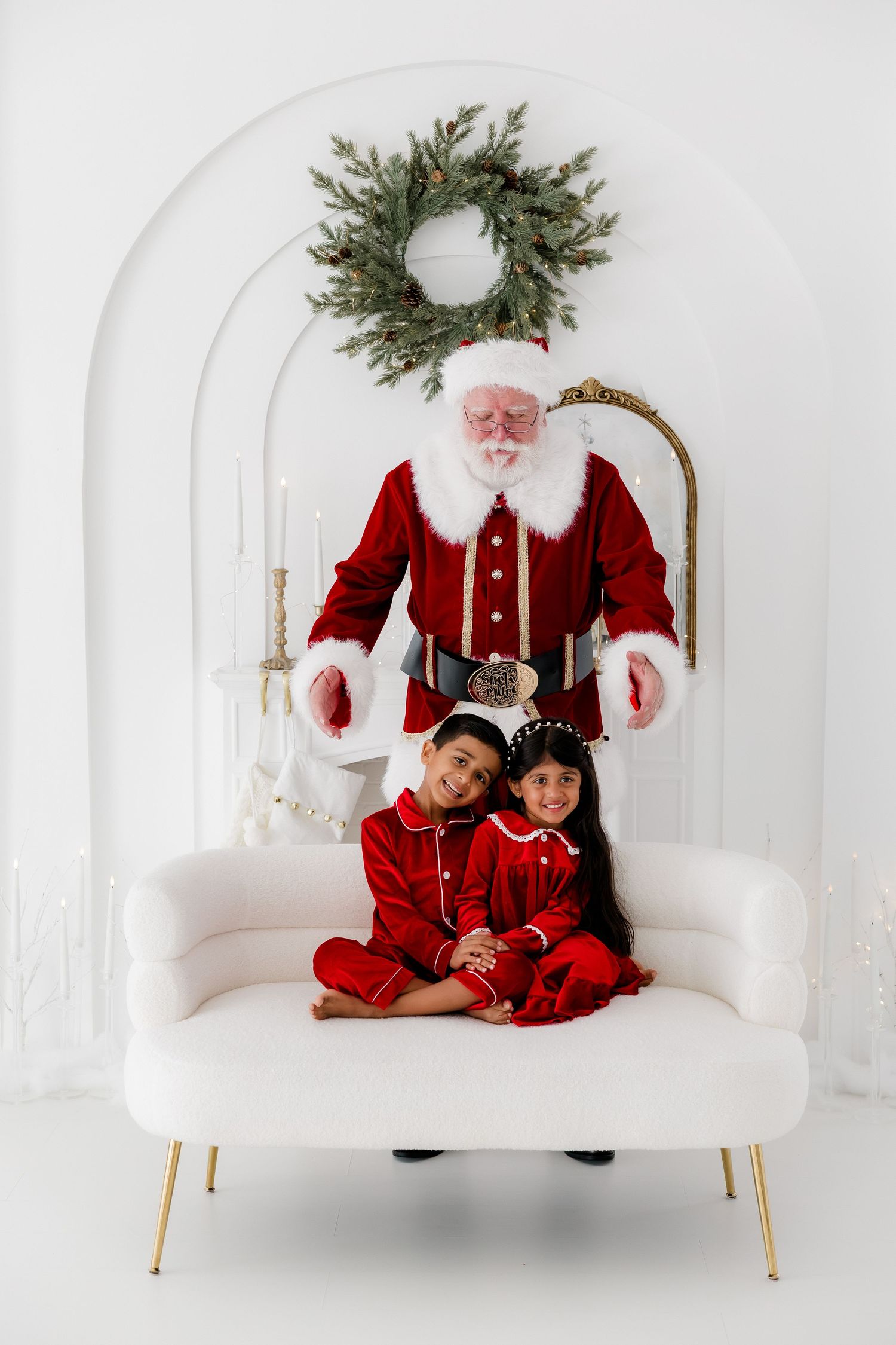Two children in red pajamas sit on a white sofa with Santa Claus behind them near a Christmas wreath.