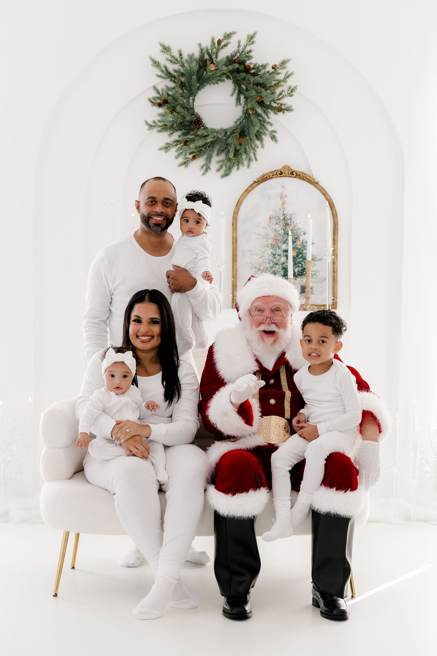 Family in white outfits poses with Santa Claus on a white sofa in a festive holiday photo studio setting.