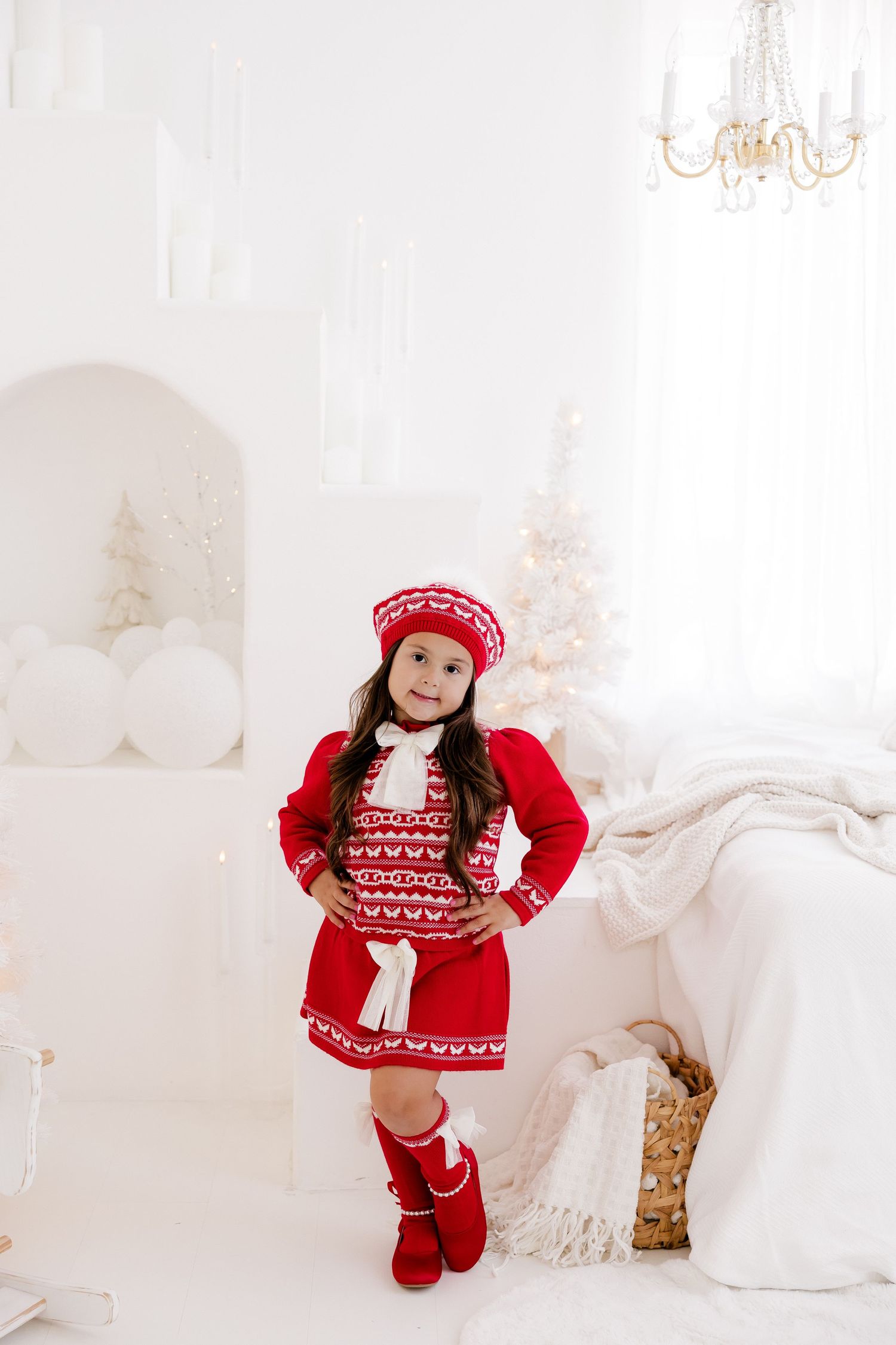 Child wearing festive red Nordic sweater poses in elegant white holiday setting with glowing decorations.