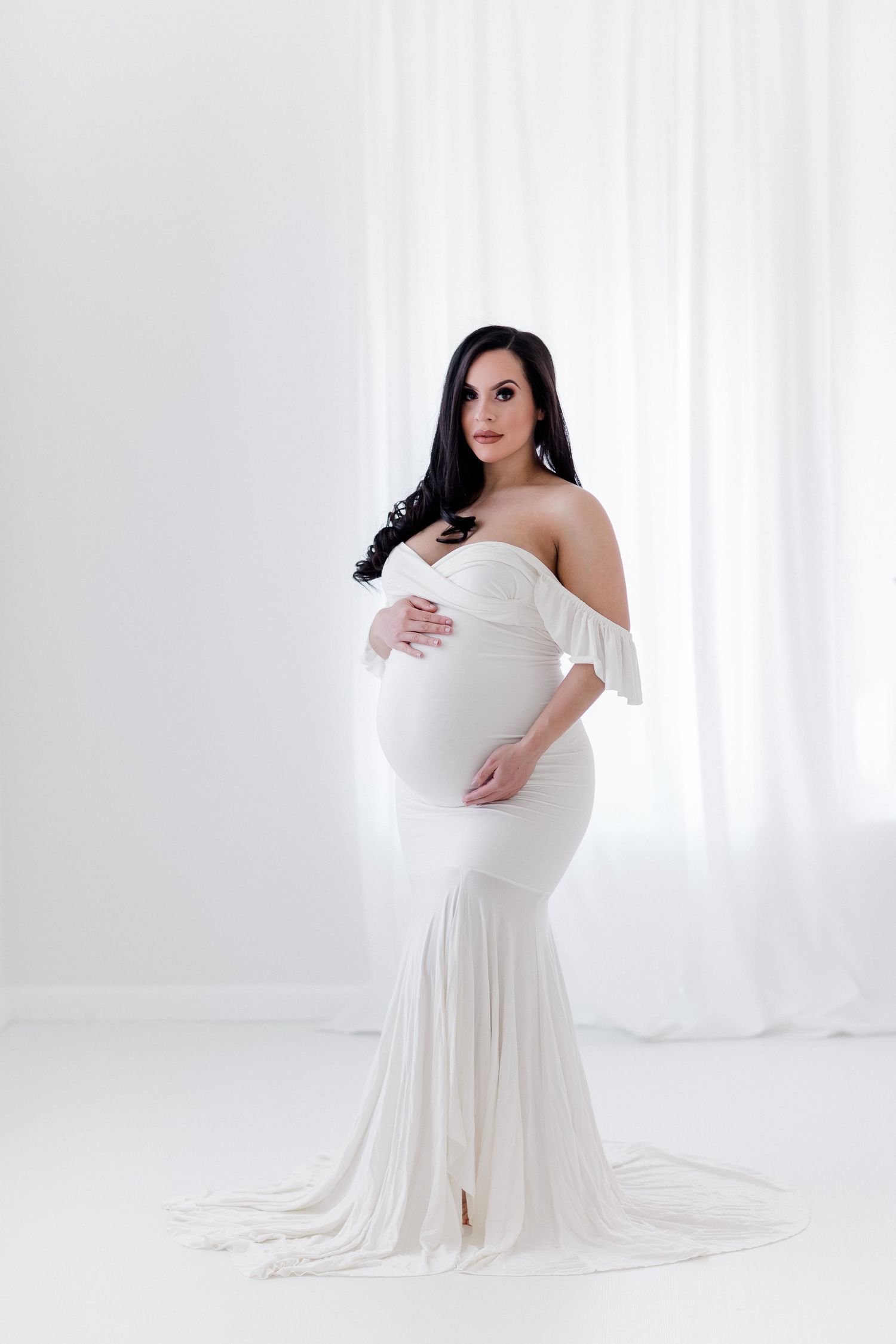 Classic maternity photo featuring white off-shoulder gown in minimalist studio.