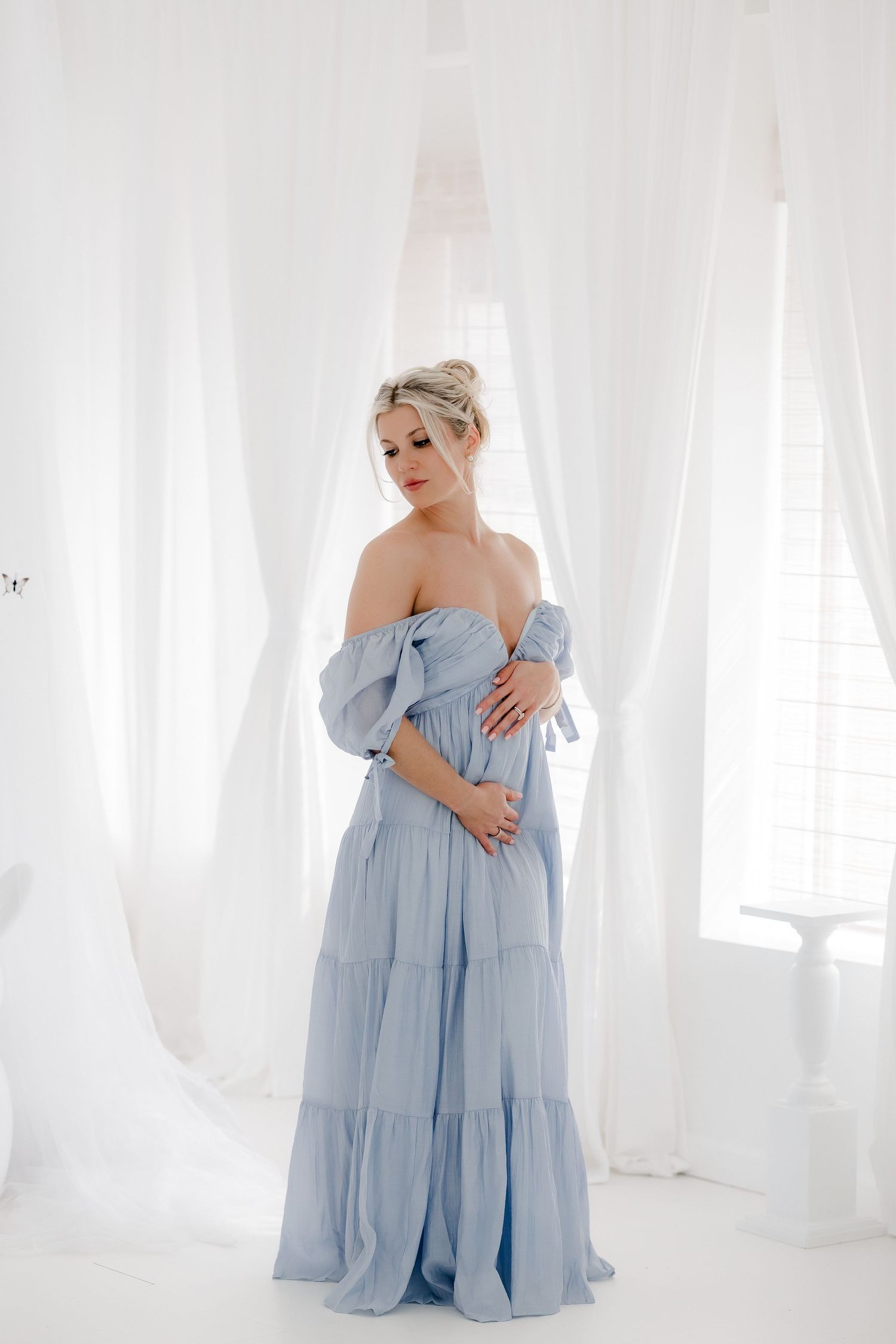 Ethereal maternity photoshoot featuring a light blue off-shoulder gown in an all-white studio setting with flowing curtains.