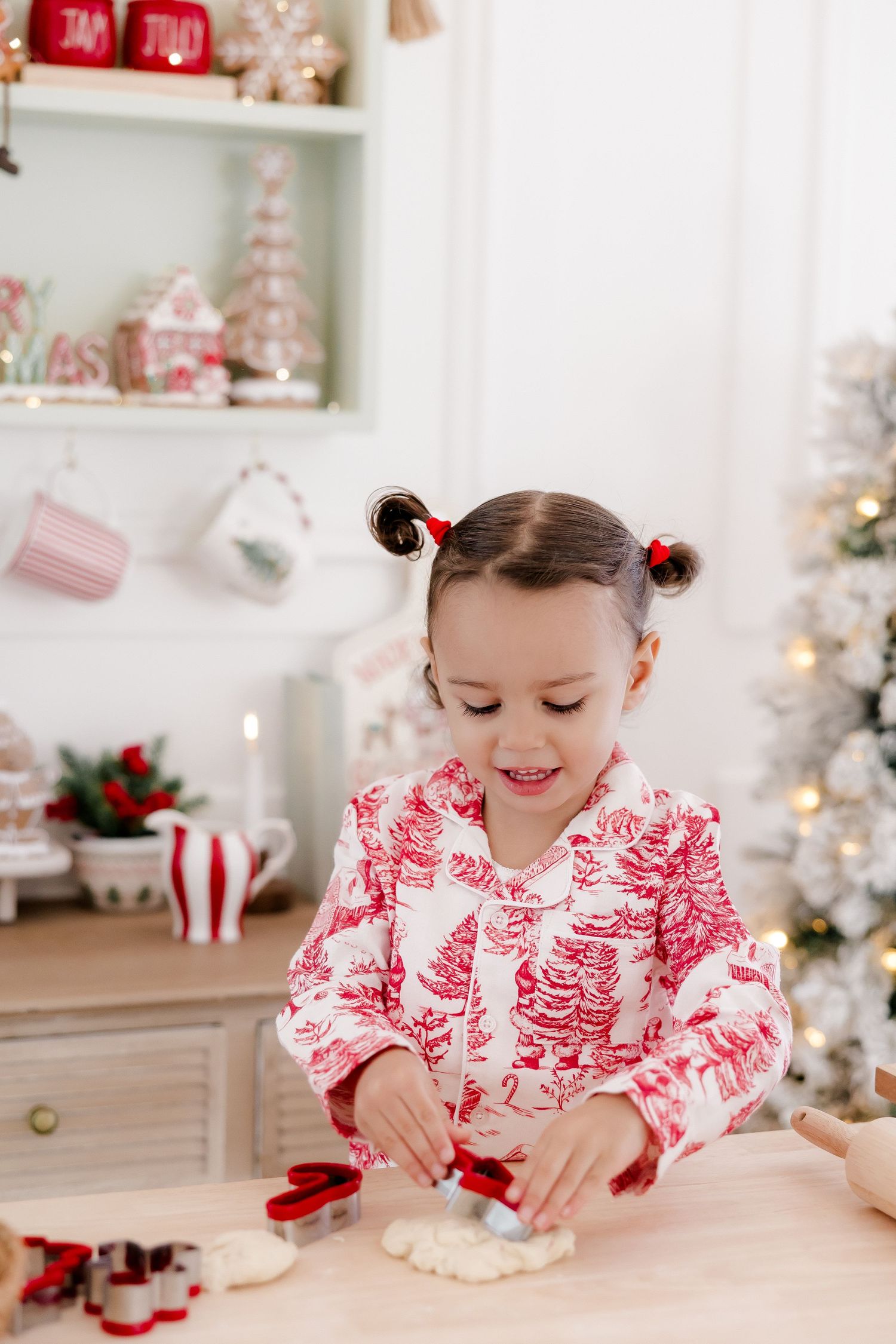 A young girl in red and white Christmas pajamas uses cookie cutters to make holiday cookies in a festive kitchen.