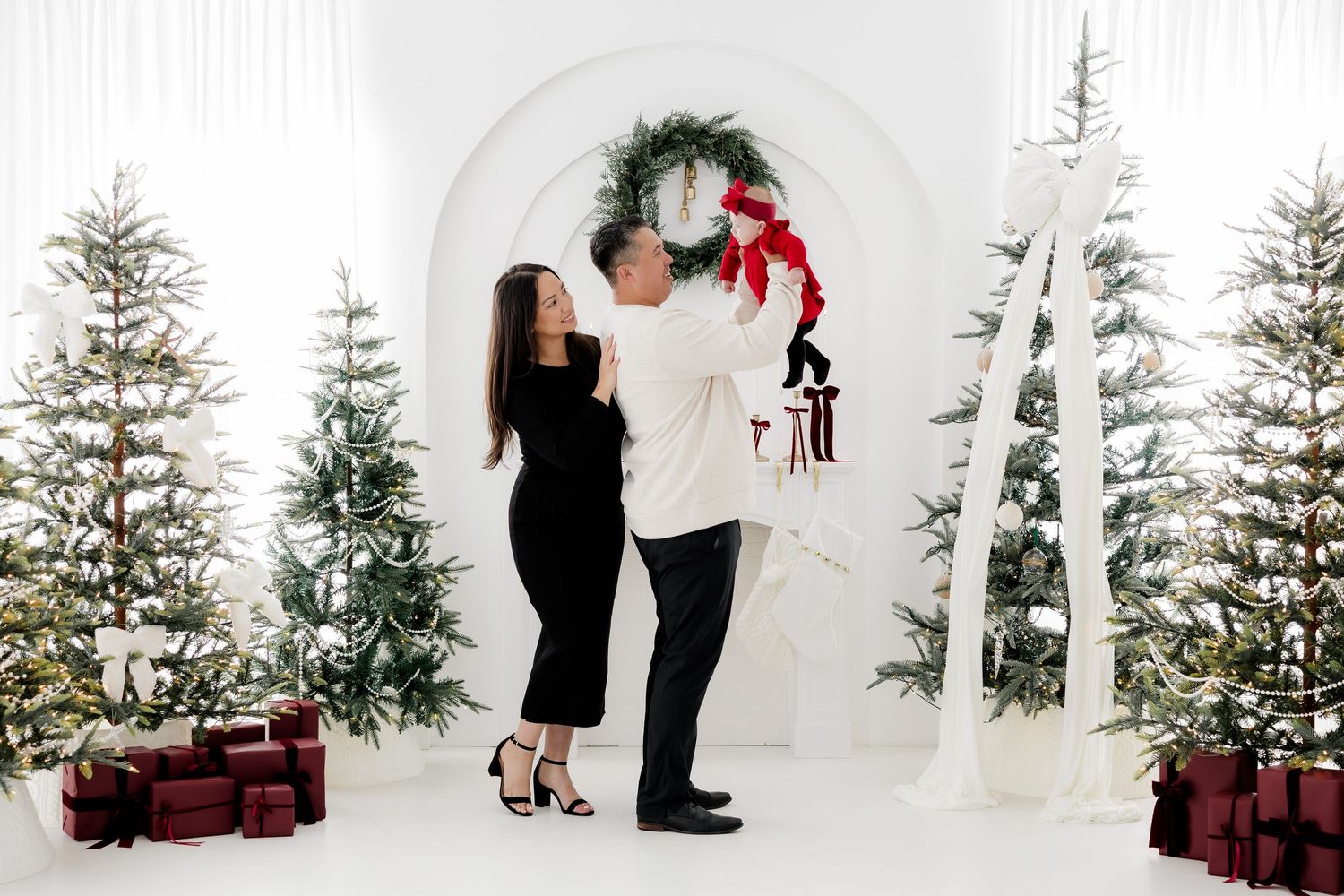 Couple poses with baby near Christmas trees and wreath arch in festive white studio holiday photo session.