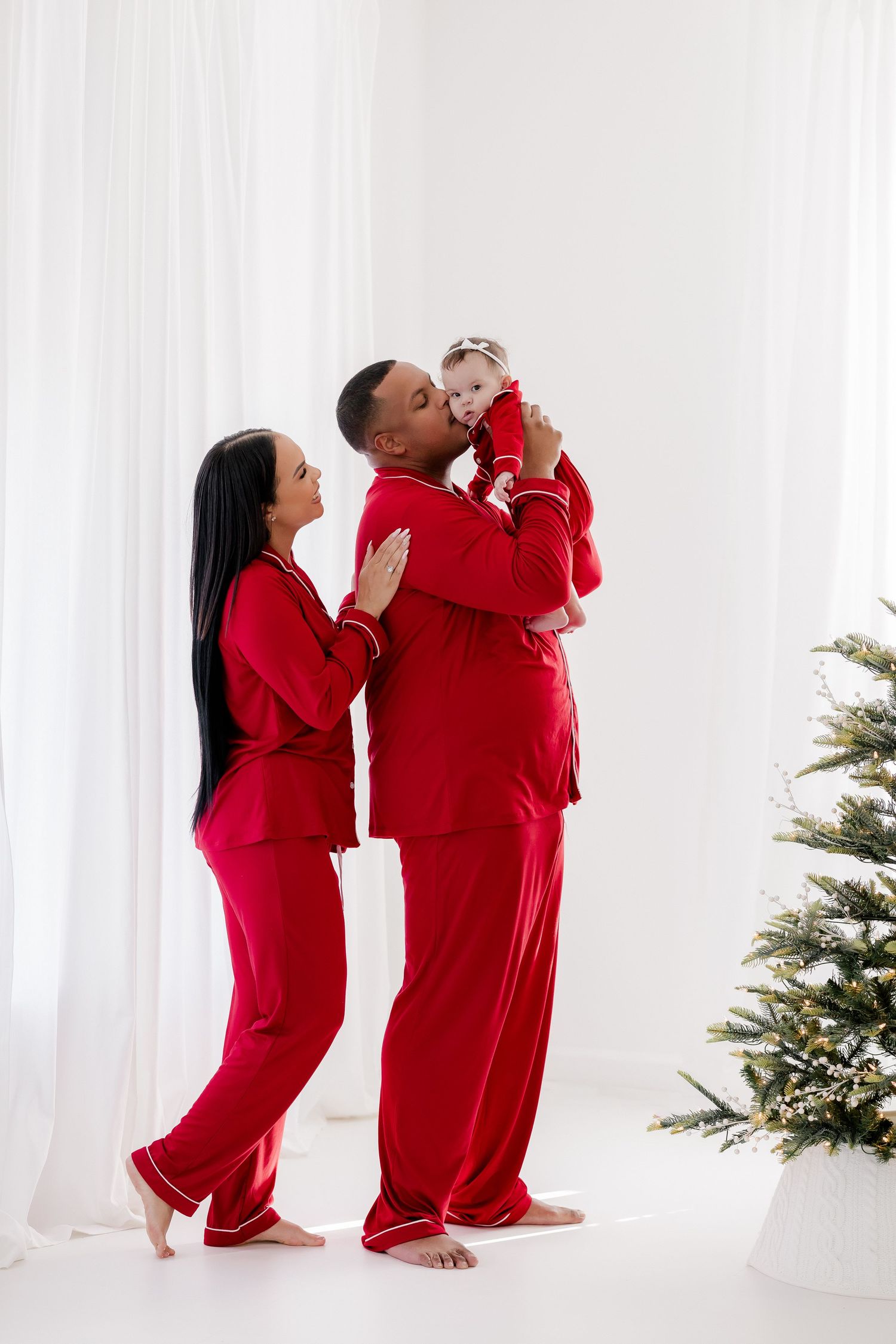 Family in matching red pajamas celebrating Christmas, father kissing baby near decorated tree.