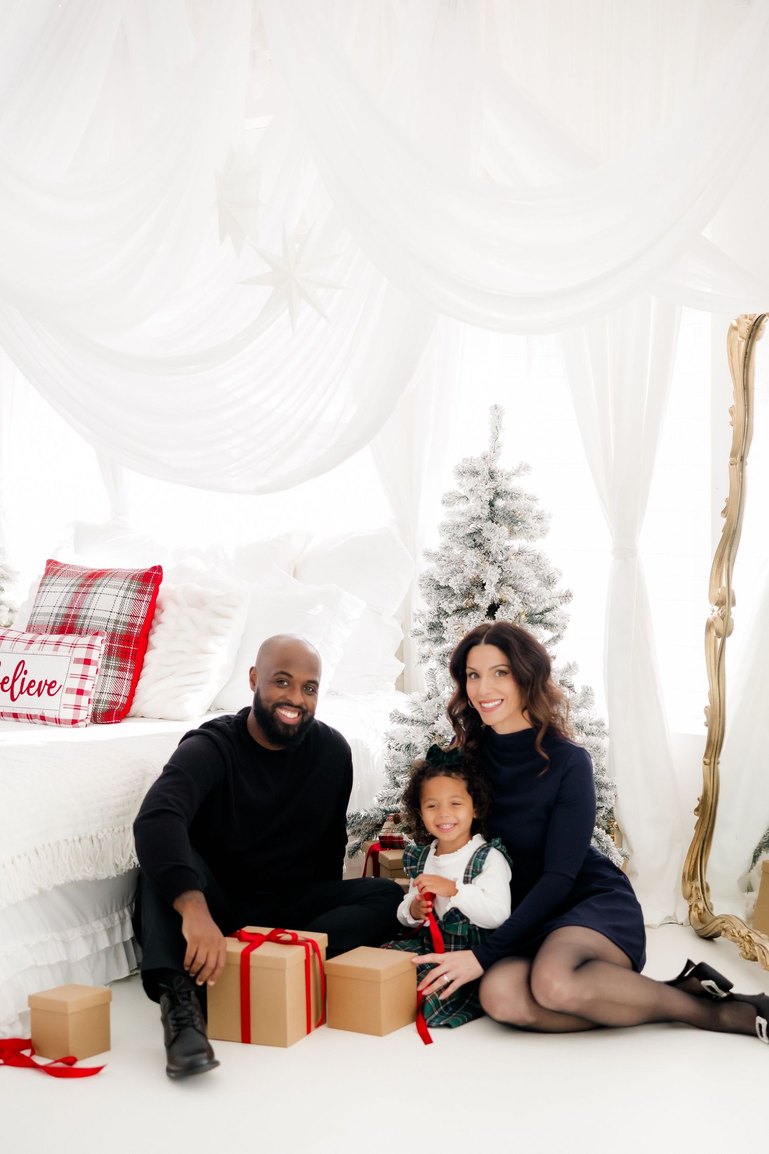 Interracial family of three posing with Christmas gifts near a flocked tree in a white holiday studio setting.