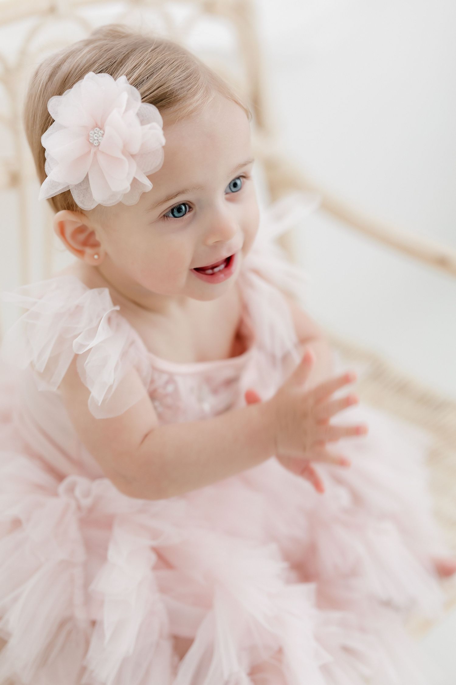 A baby wearing a delicate pink tulle dress and flower headband smiles and plays in a bright white studio setting.