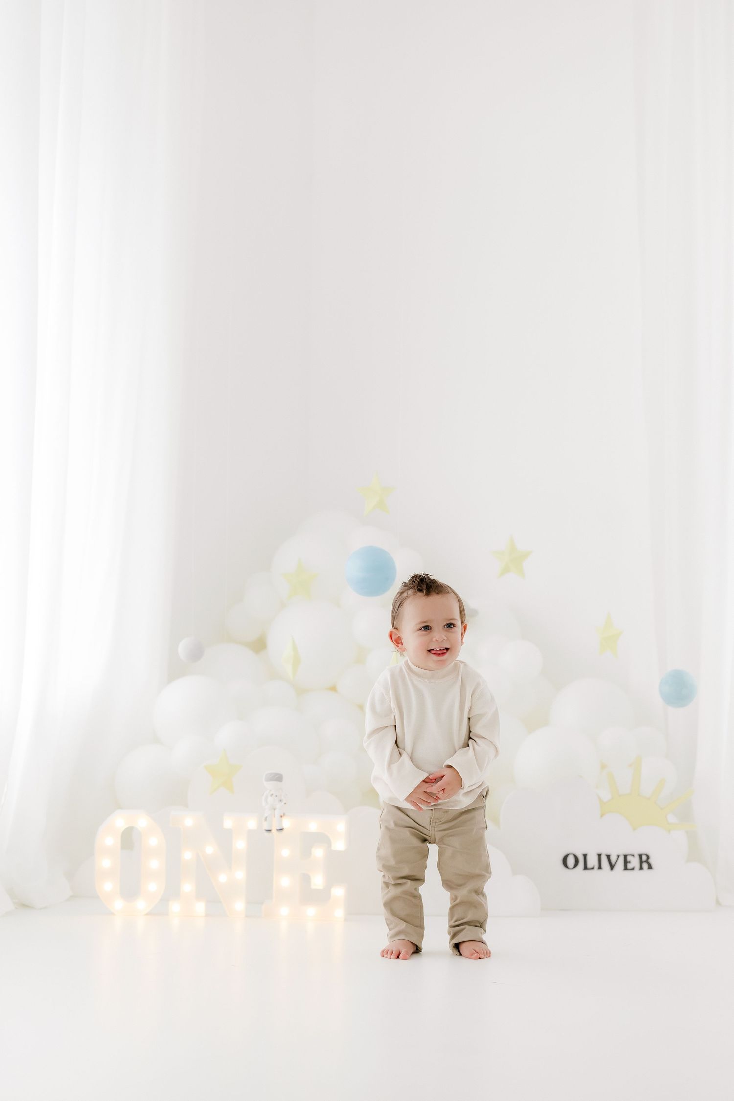 Sweet baby in beige outfit plays on white backdrop with pastel balloons and star decorations in dreamy nursery setting.