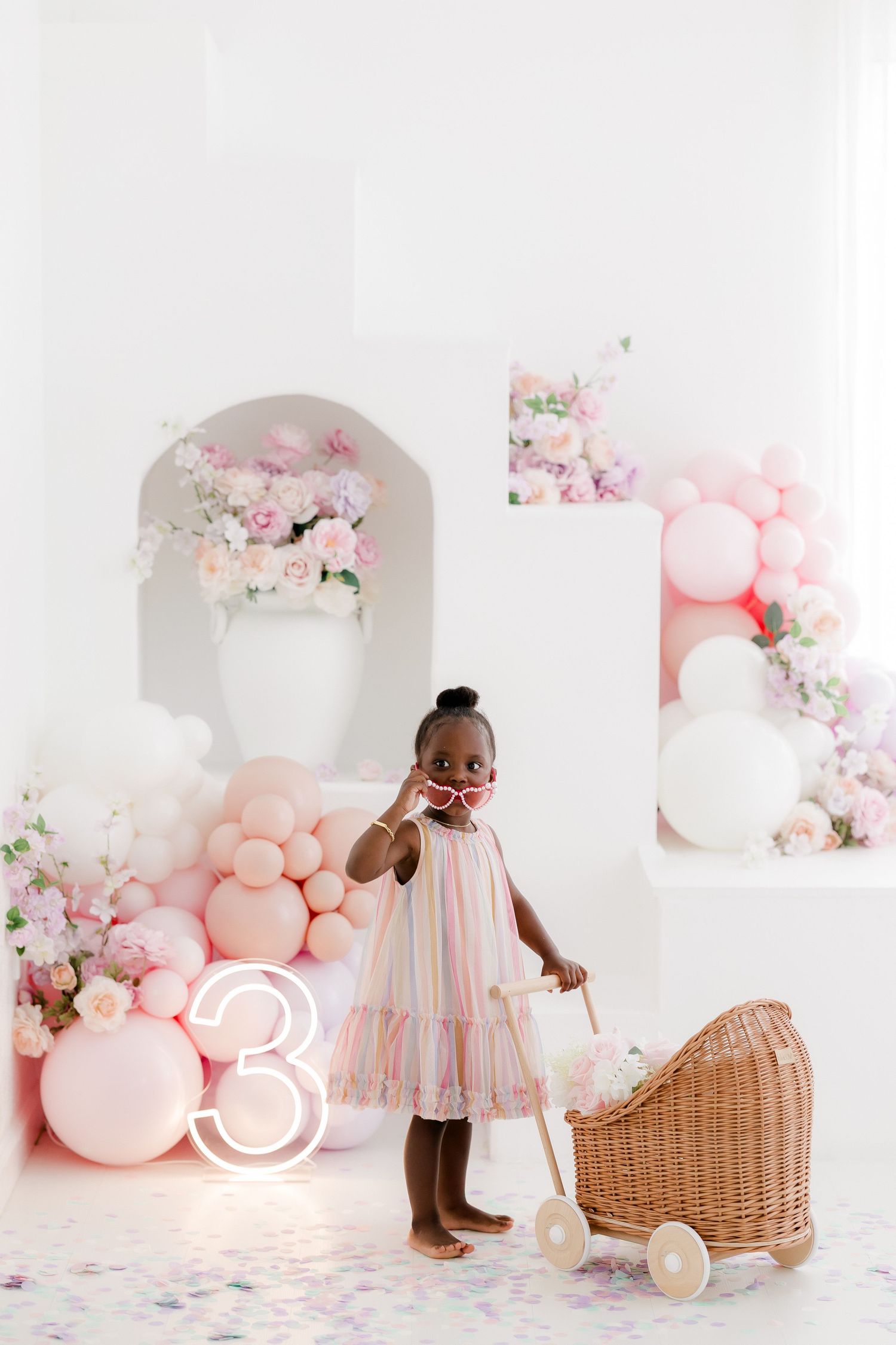 A young girl in a pastel dress poses by a wicker pram at a pink balloon 3rd birthday party setup.