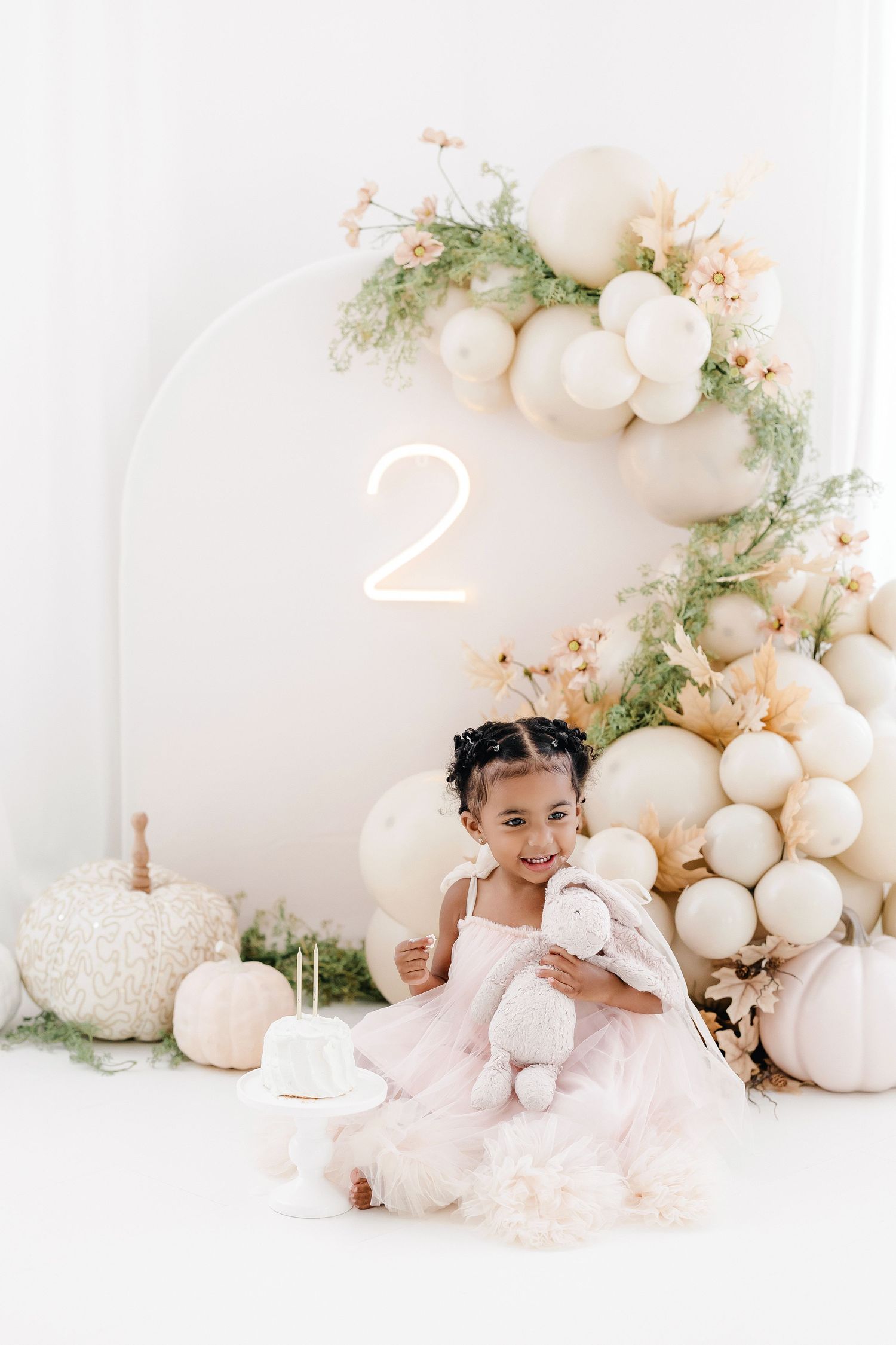 Elegant white balloon arch with delicate greenery and soft pink accents creates a dreamy cake smash photo backdrop.