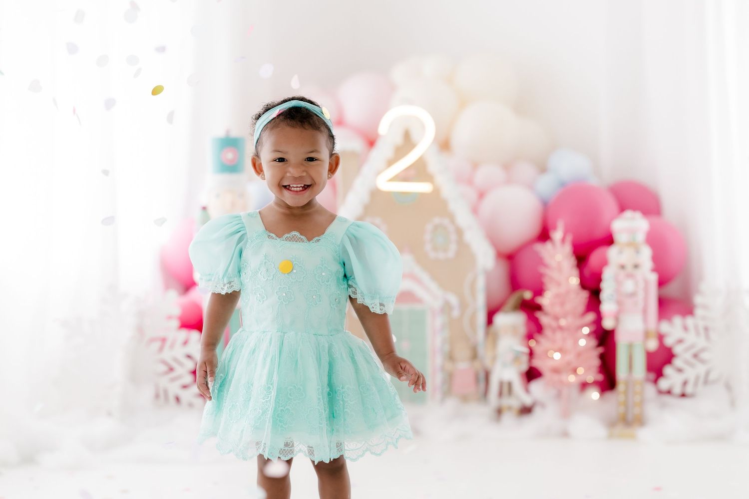 A toddler in a mint green tulle dress poses and plays in front of a pink and white party backdrop.