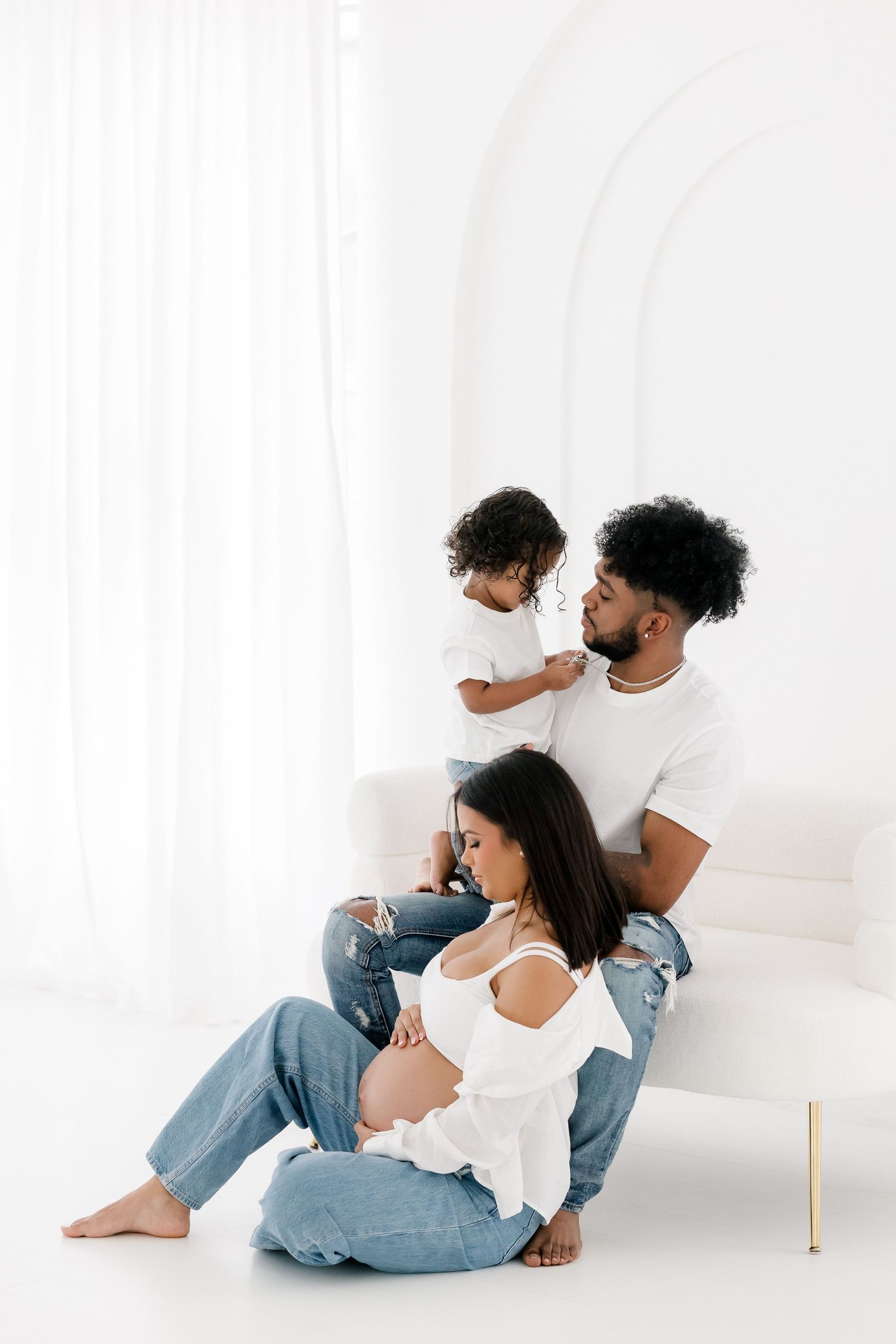 Couple in white tops and jeans cuddle together in minimalist white studio setting during intimate photoshoot.