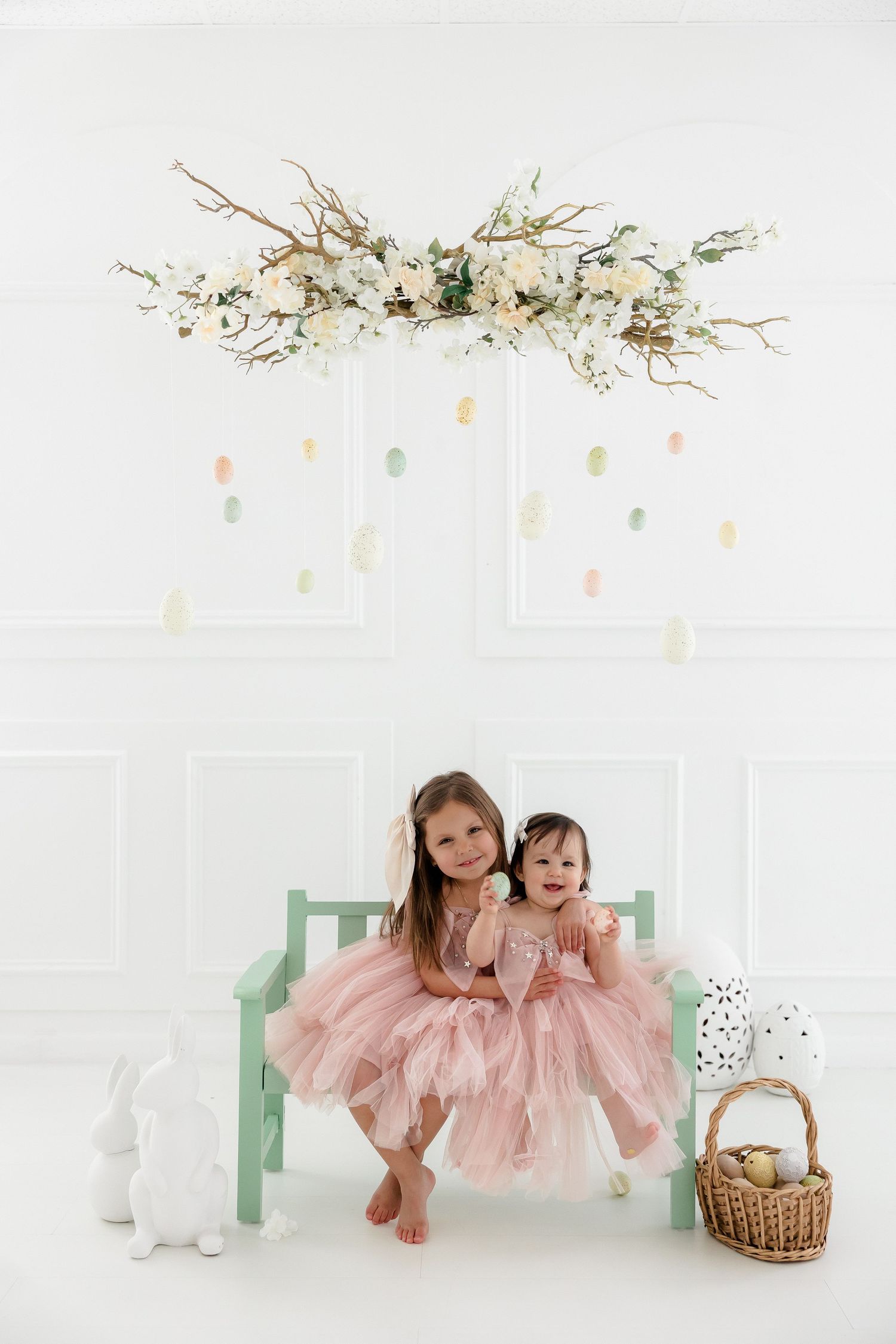 Children in blush pink tulle dresses pose in a bright white studio setting with delicate floral decorations.