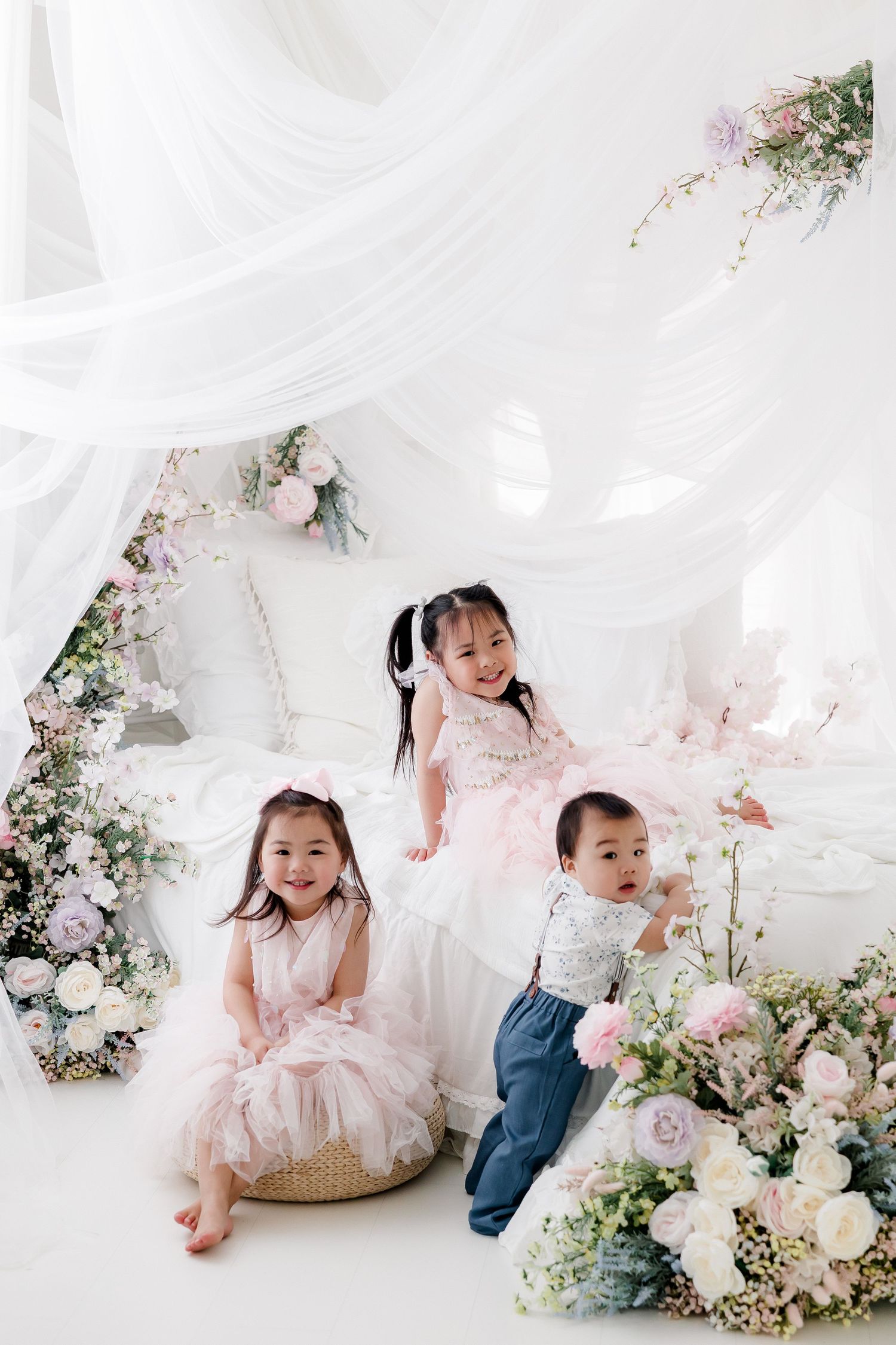Children playing together in a dreamy white studio setting with floral decorations and flowing fabric.