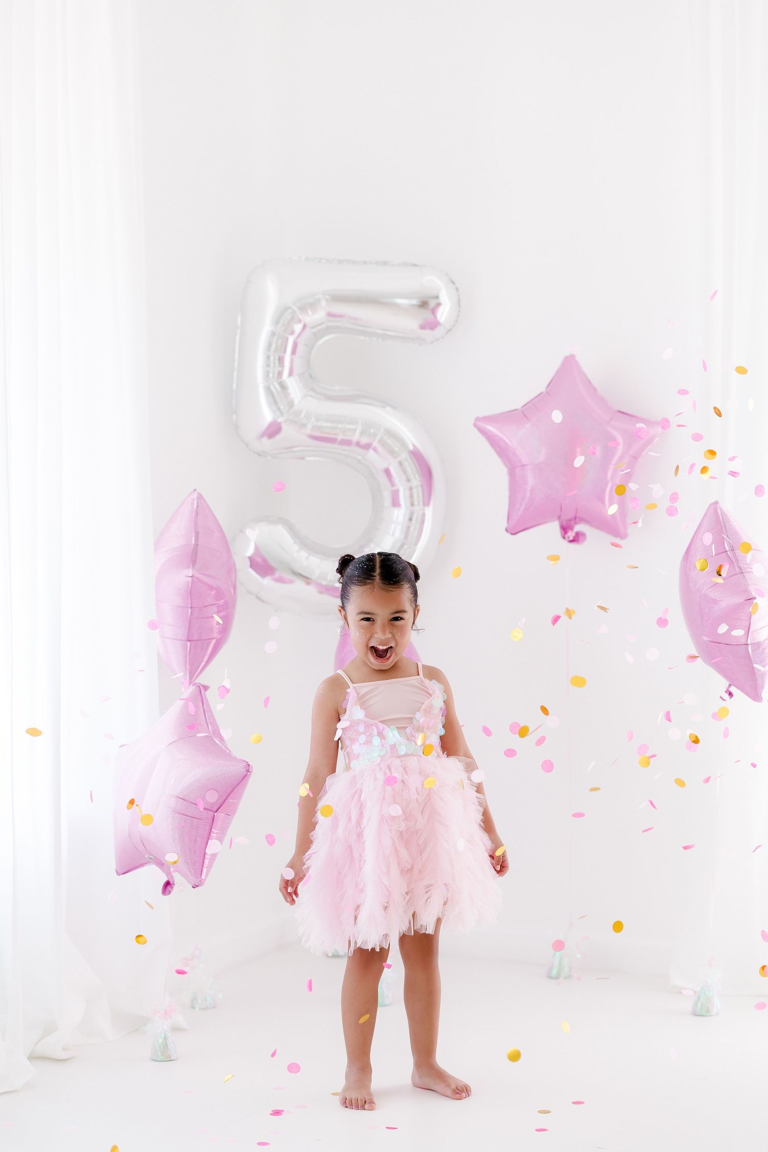 Pink and white themed first birthday photoshoot setup with star balloons, confetti and tulle dress decorations.