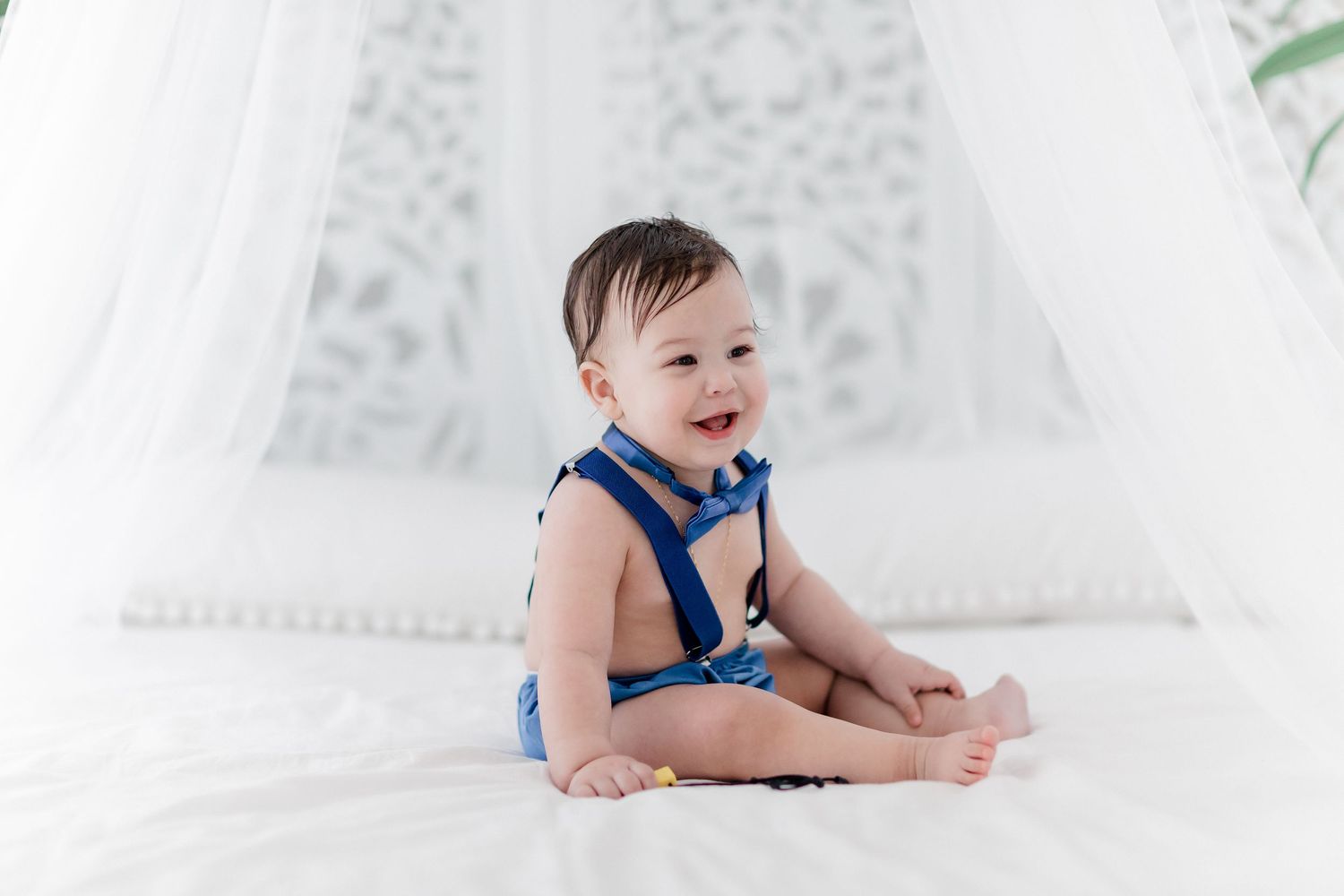 A baby in a blue outfit lies on white fabric while smiling and playing during a photo session.