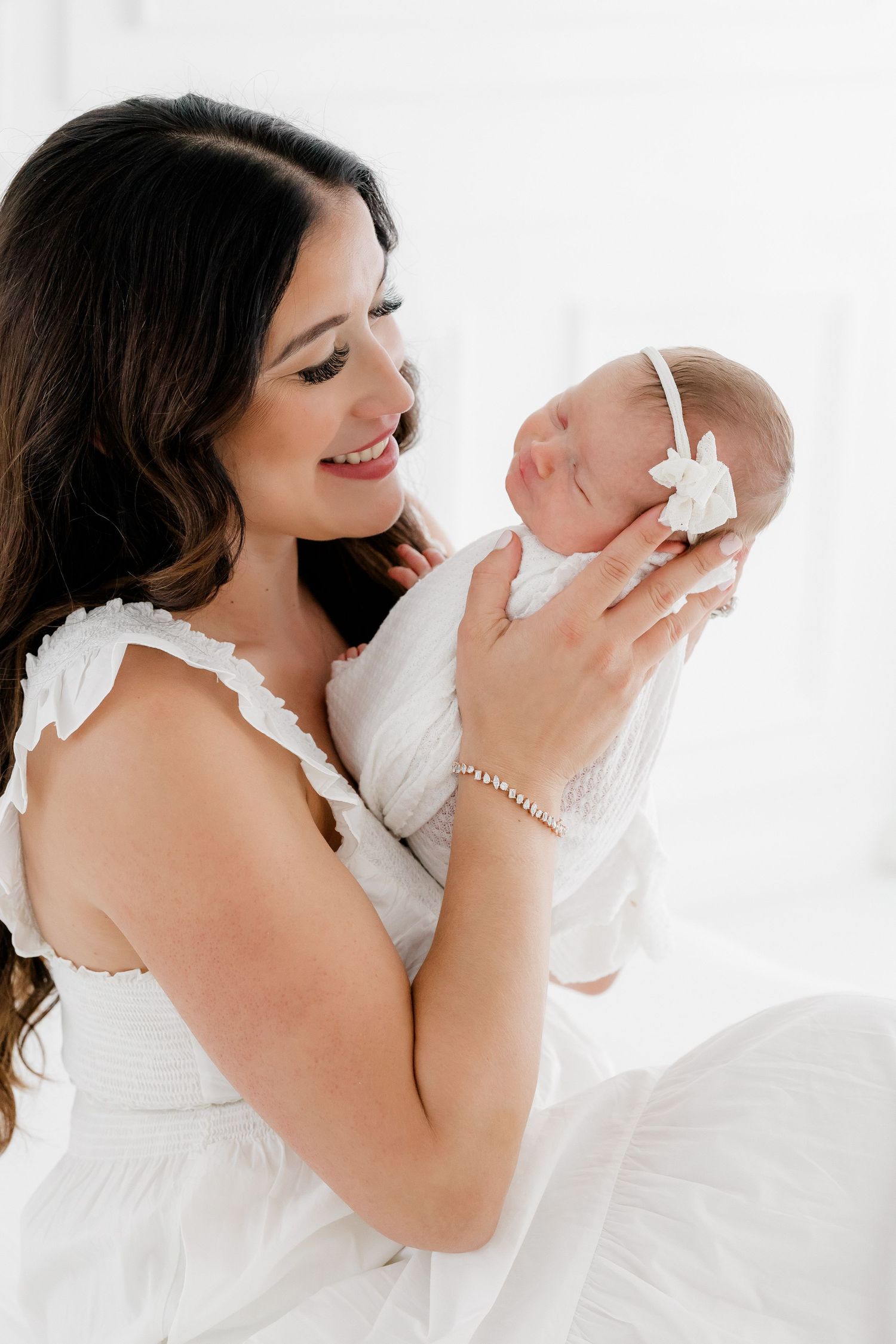 Mother in white dress holding newborn against bright studio backdrop in loving embrace.