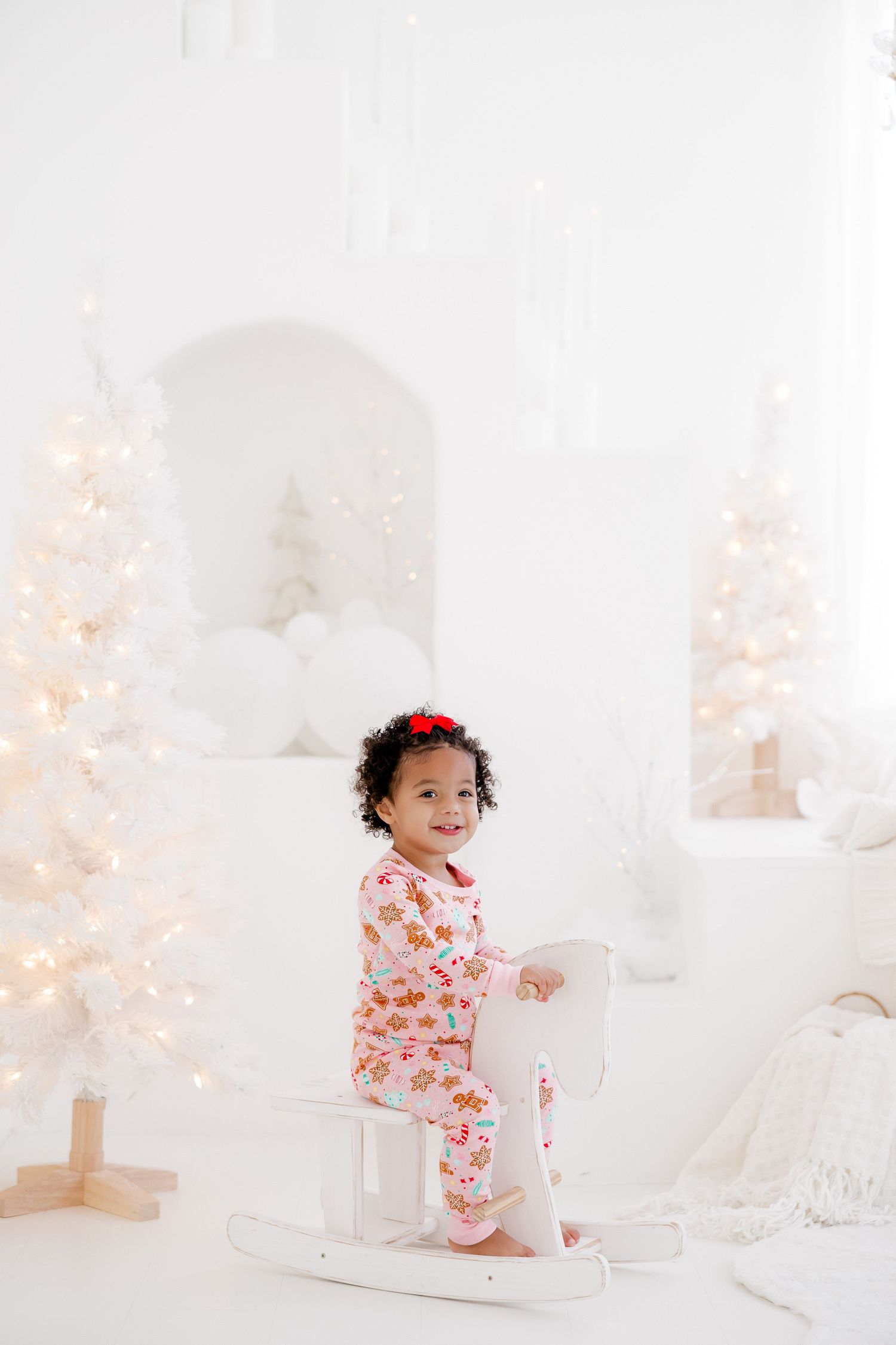 Small child in floral pajamas sits on a rocking horse surrounded by illuminated white Christmas trees.