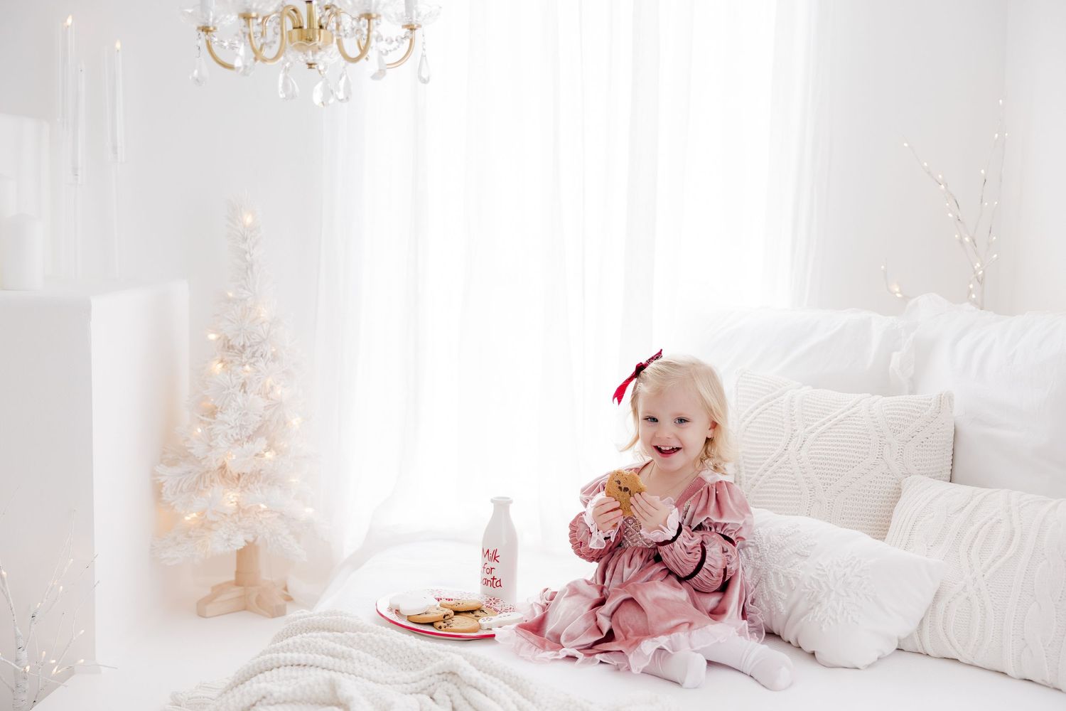 Baby in pink pajamas playing on white bedding with Christmas decor in background.nj holiday mini sessions